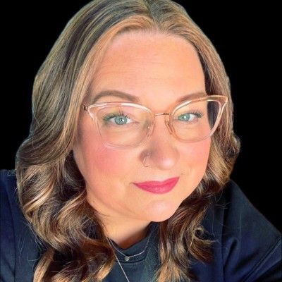 Close-up portrait of a smiling person with wavy hair, glasses, and a dark top on a black background