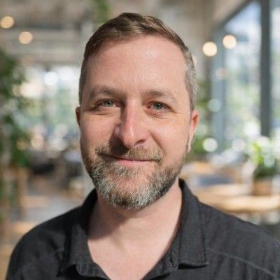 Smiling man in a dark shirt, seated indoors with a blurred café-style background