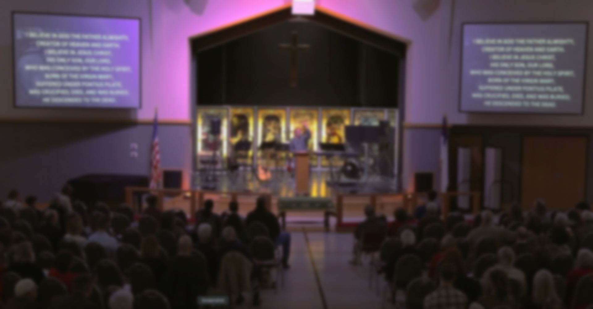 A man is standing at a podium with his arms outstretched in front of a stained glass window.