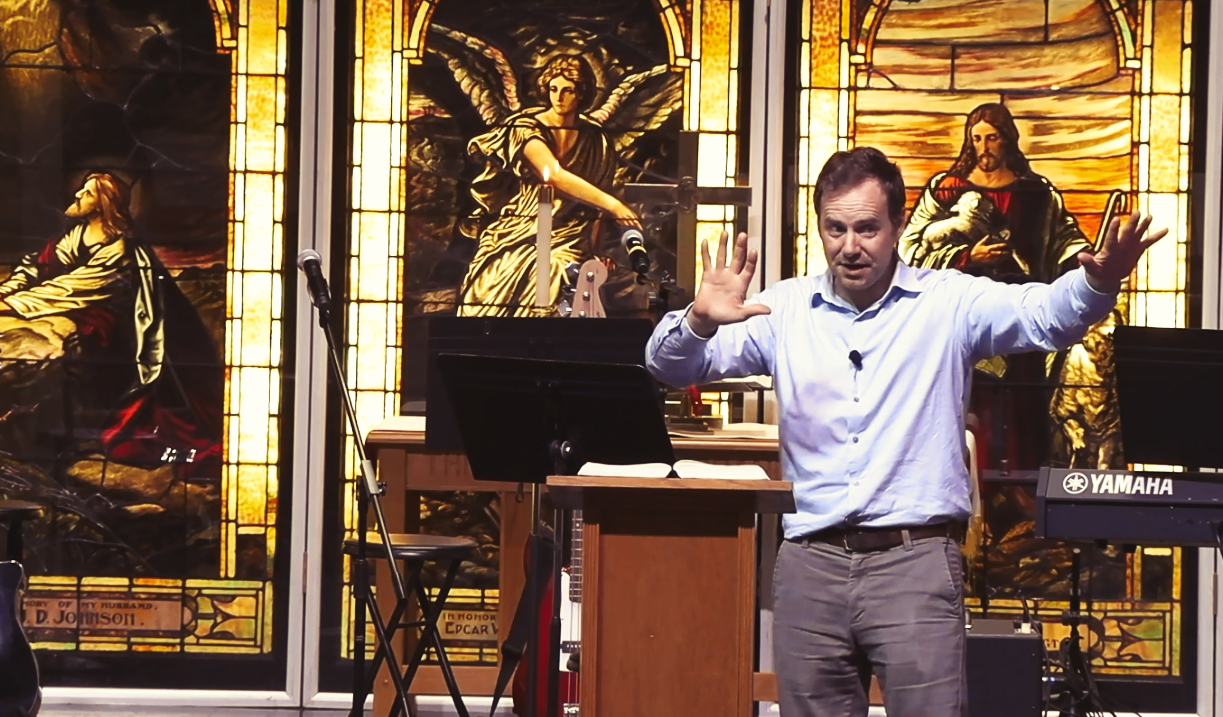 A man is standing at a podium with his arms outstretched in front of a stained glass window.