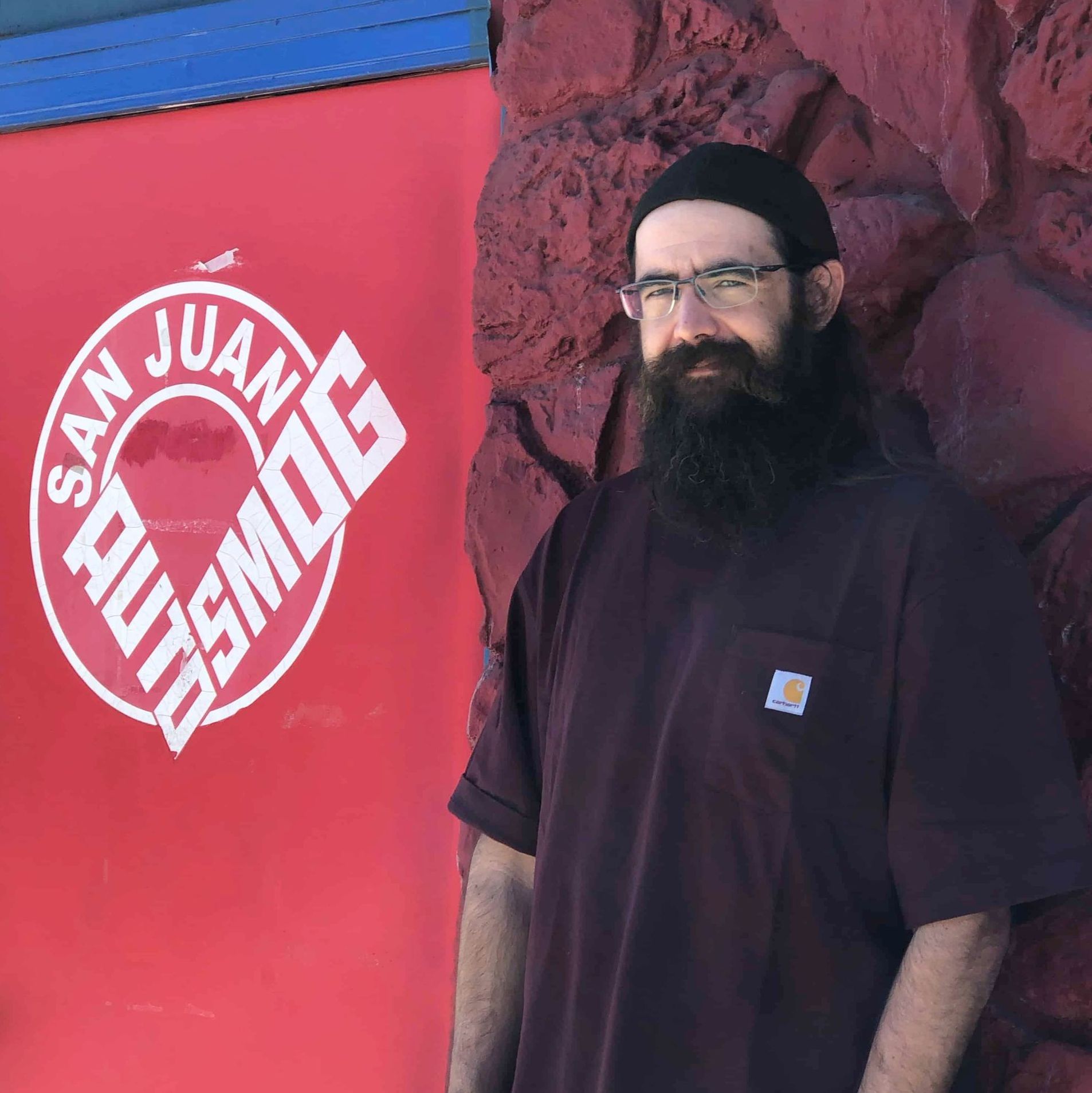 A man with a beard is standing in front of a red sign that says san juan auto smog