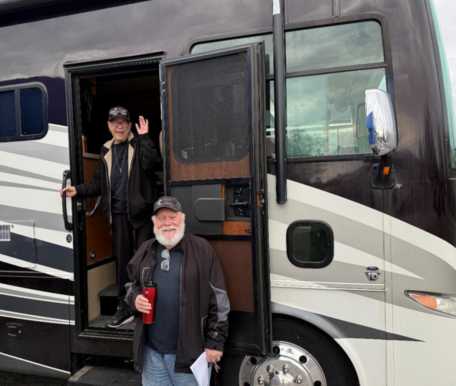 Two people wave and smile beside a large black and white RV, one holding a red cup.