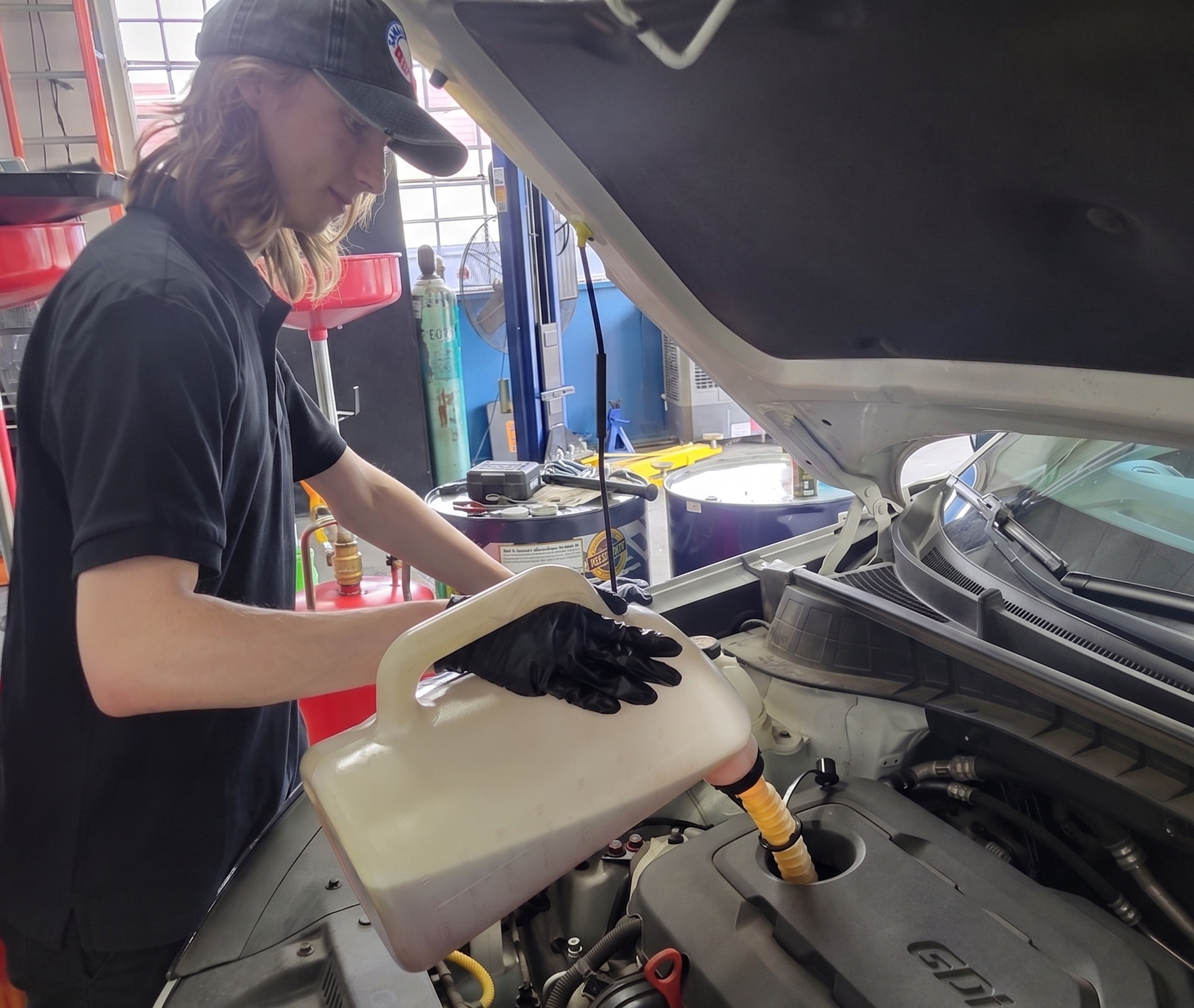 Mechanic pouring fluid into a car engine bay with the hood open in a garage