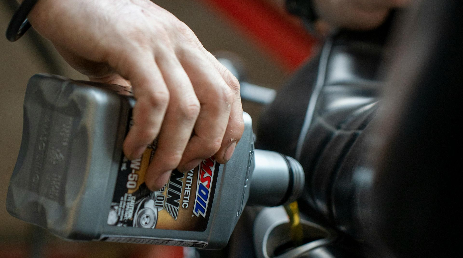 A mechanic wearing black gloves pours motor oil from a grey plastic bottle into an open car engine.