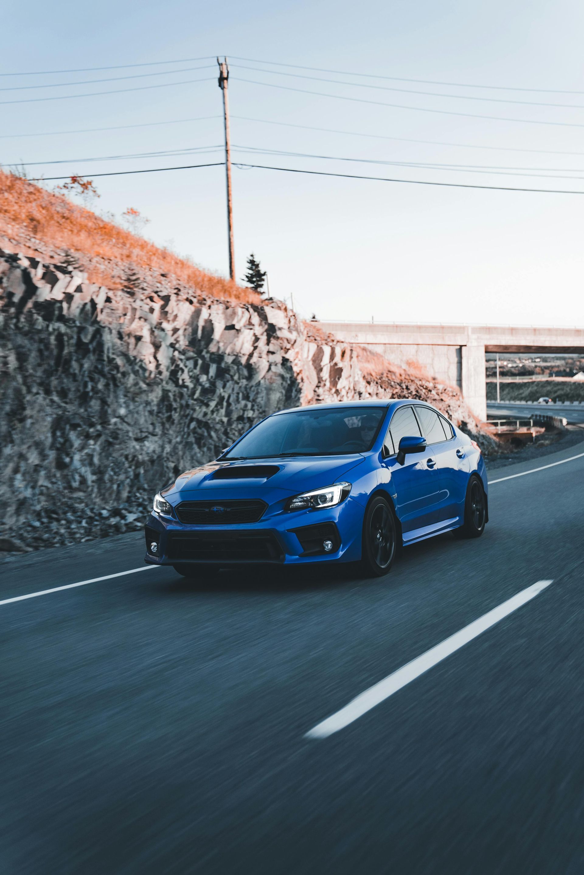 Blue sports car driving on a road beside a stone wall and bridge under a clear sky