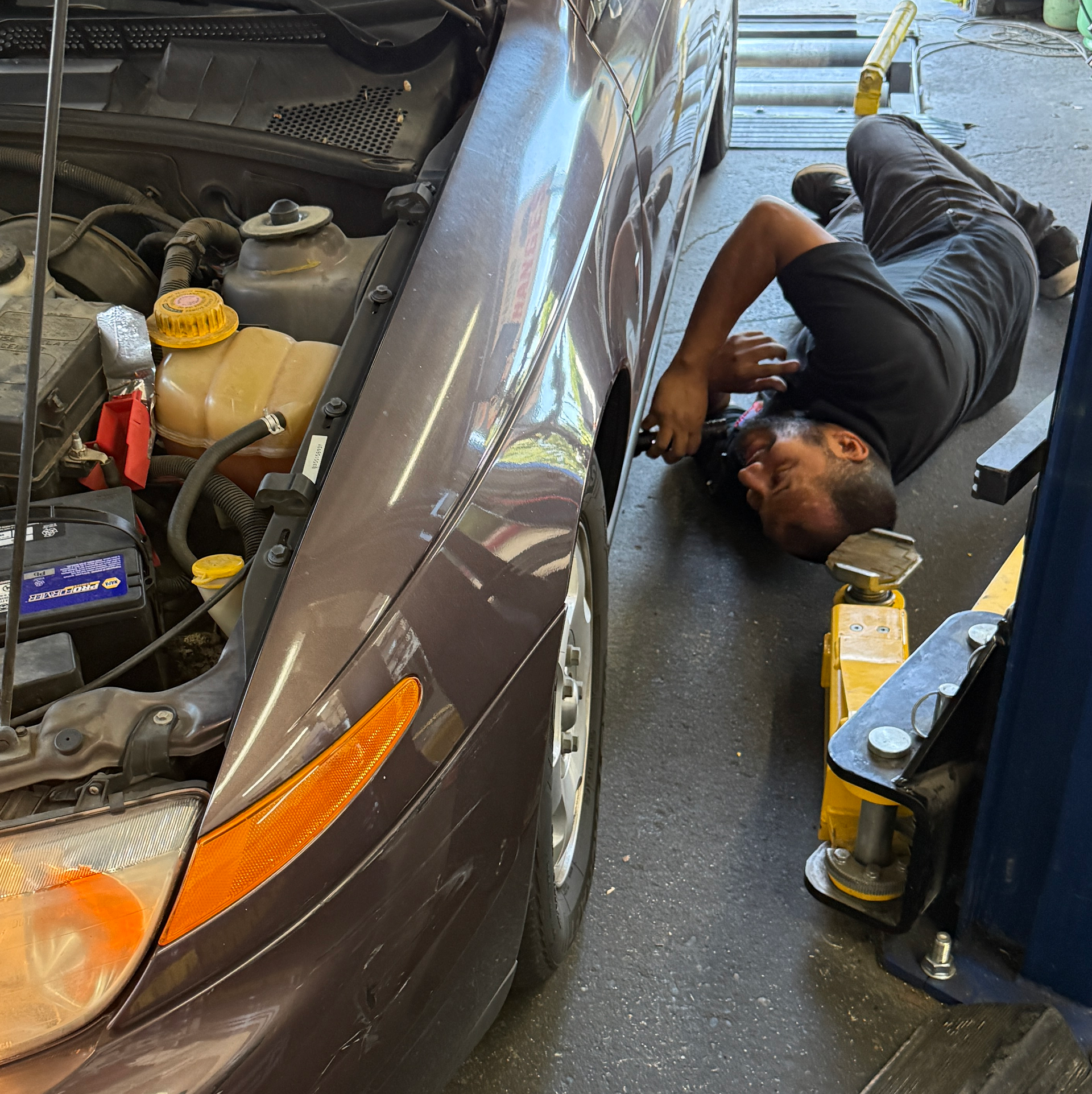 Mechanic lying beside a brown car, working near the front wheel inside an auto shop.