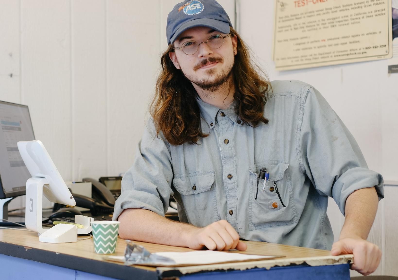 Man in denim shirt and blue cap sitting at a desk, speaking in an office