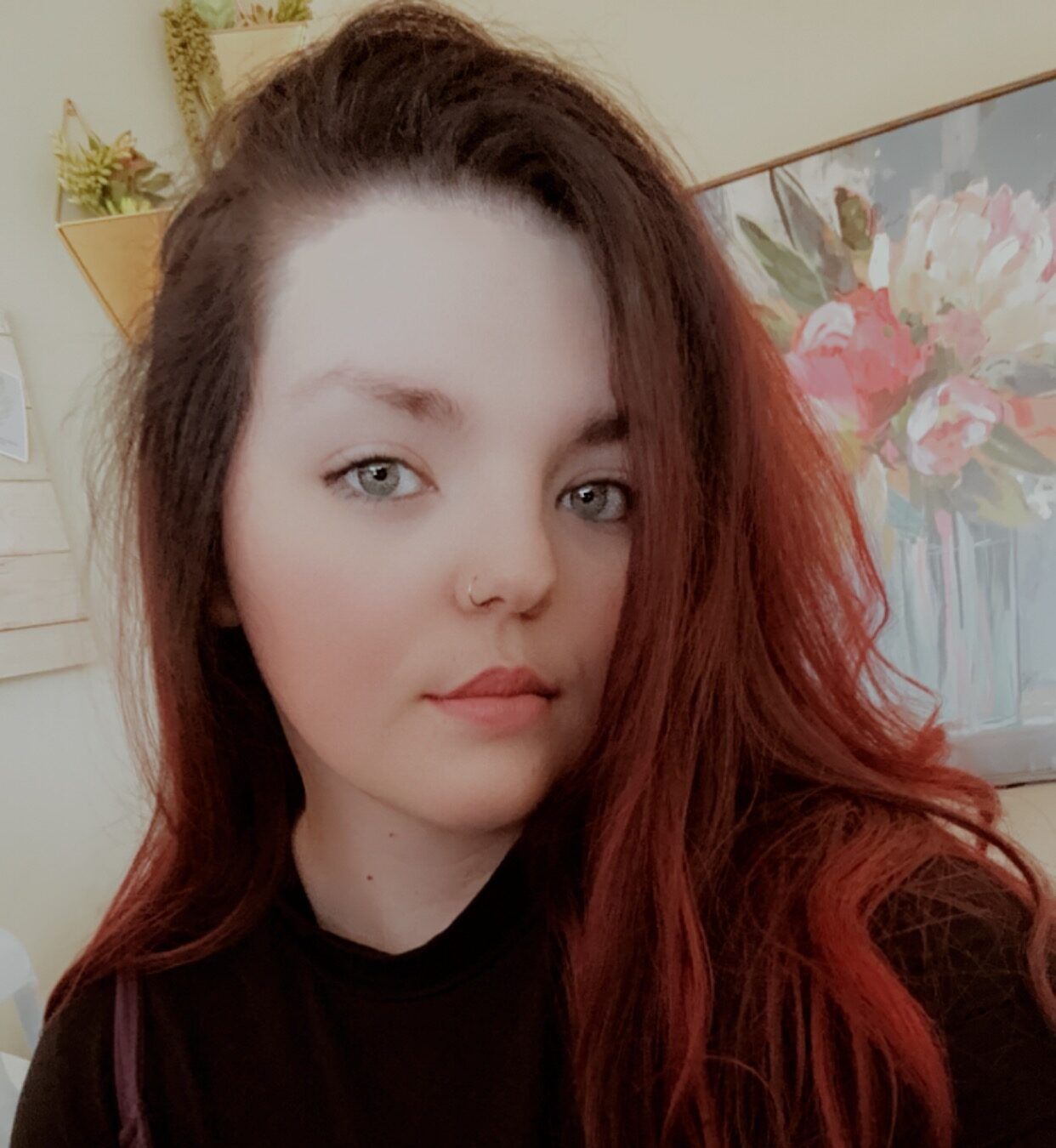 A woman with long red hair is taking a selfie in front of a painting of flowers.
