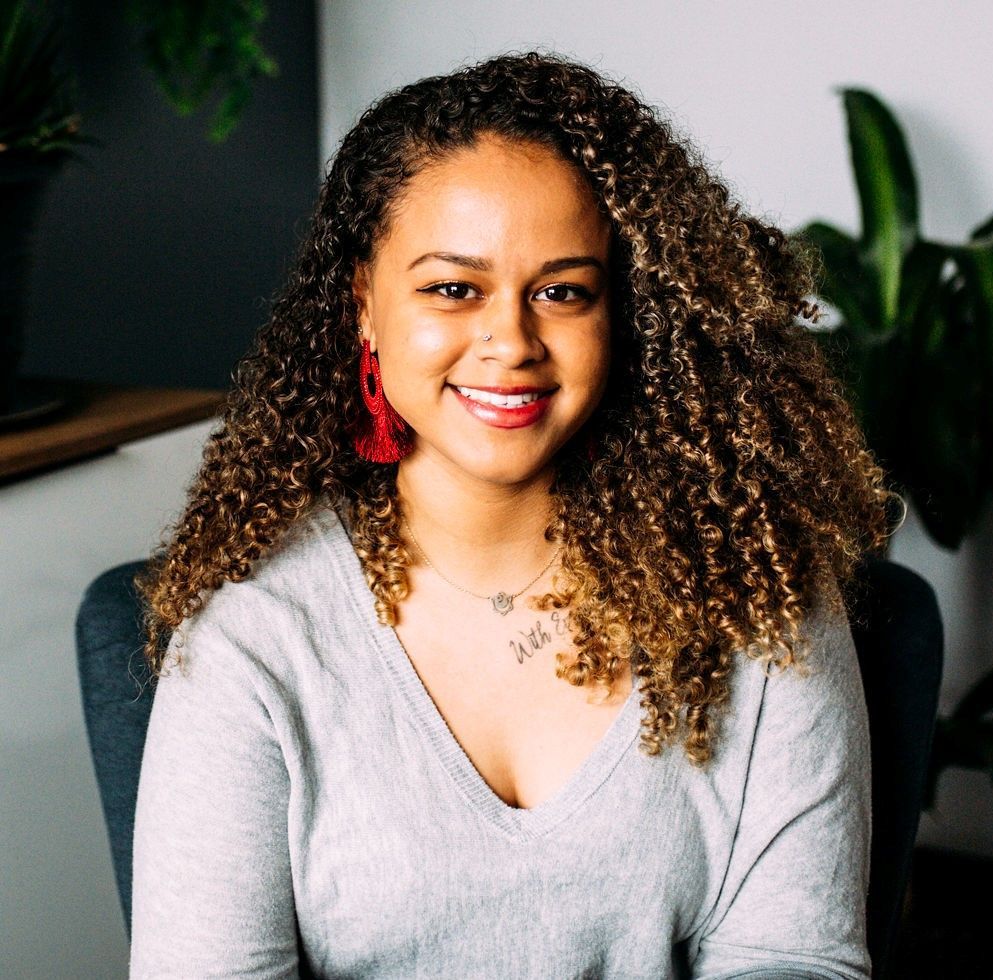 A woman with curly hair is sitting in a chair and smiling for the camera.
