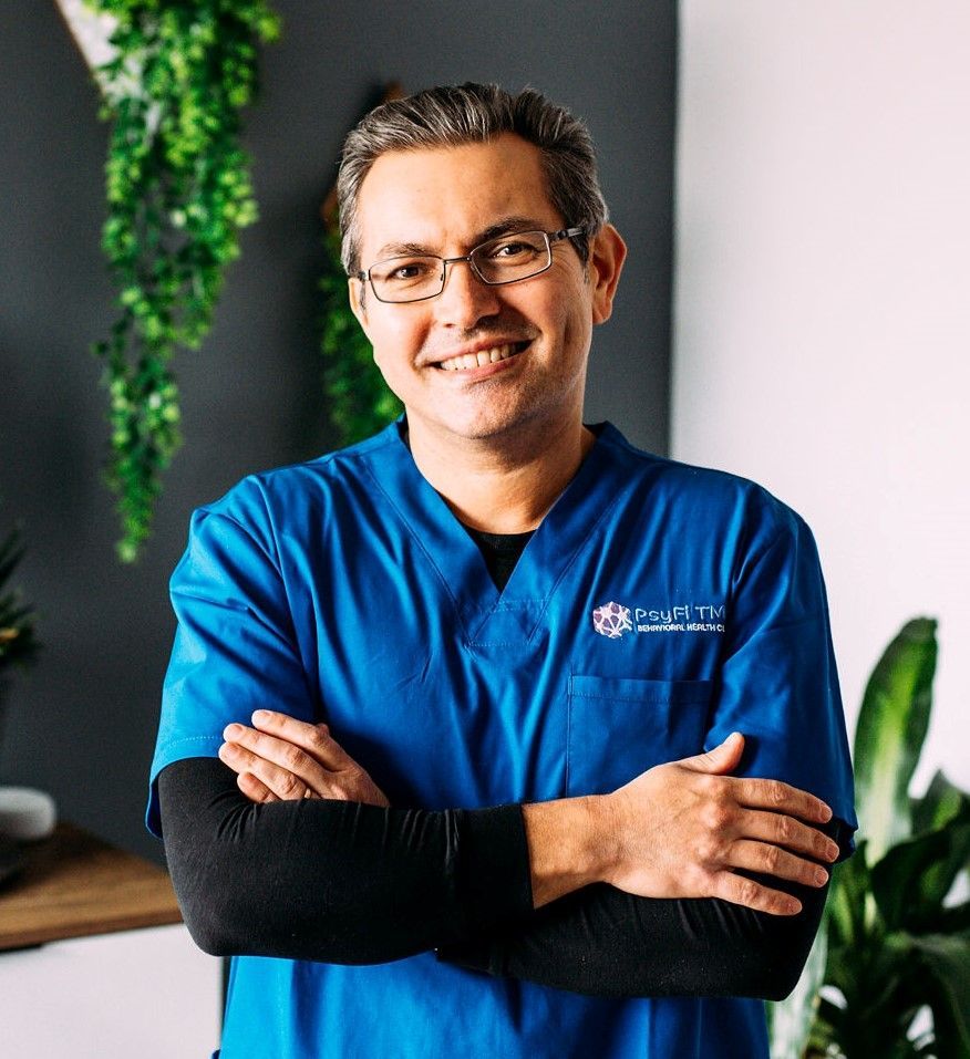 A man in a blue scrub is standing with his arms crossed and smiling.