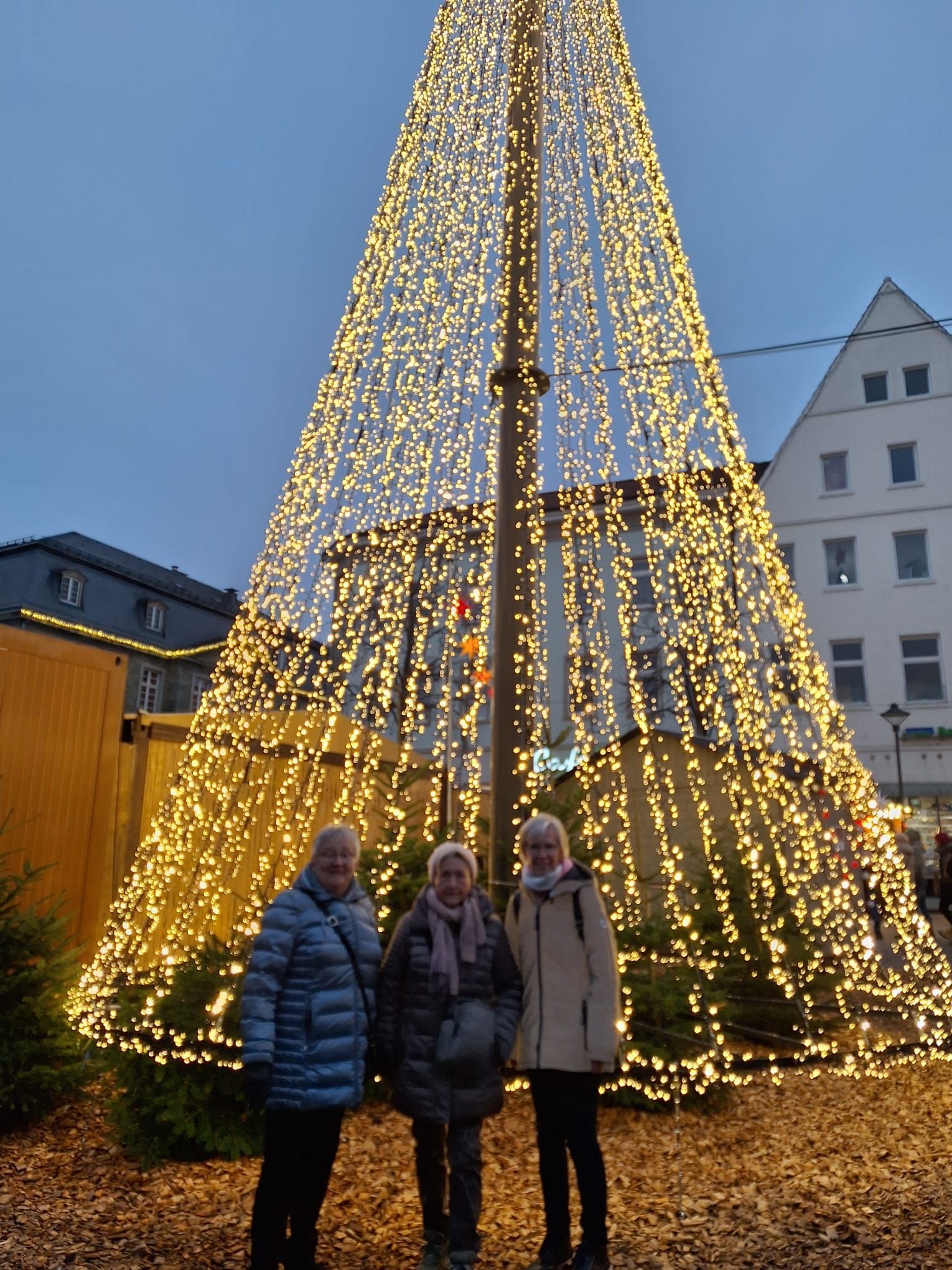 Drei Frauen stehen vor einem großen, mit Lichtern geschmückten Weihnachtsbaum.