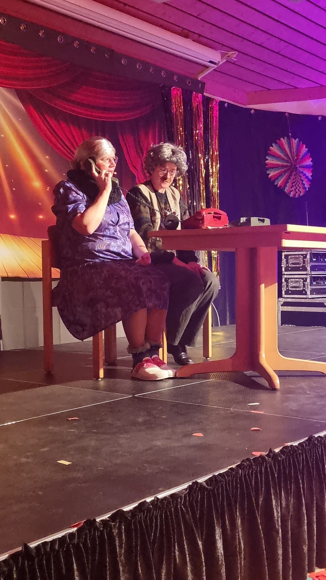 Zwei Frauen sitzen an einem Tisch auf einer Bühne und telefonieren mit ihren Handys.