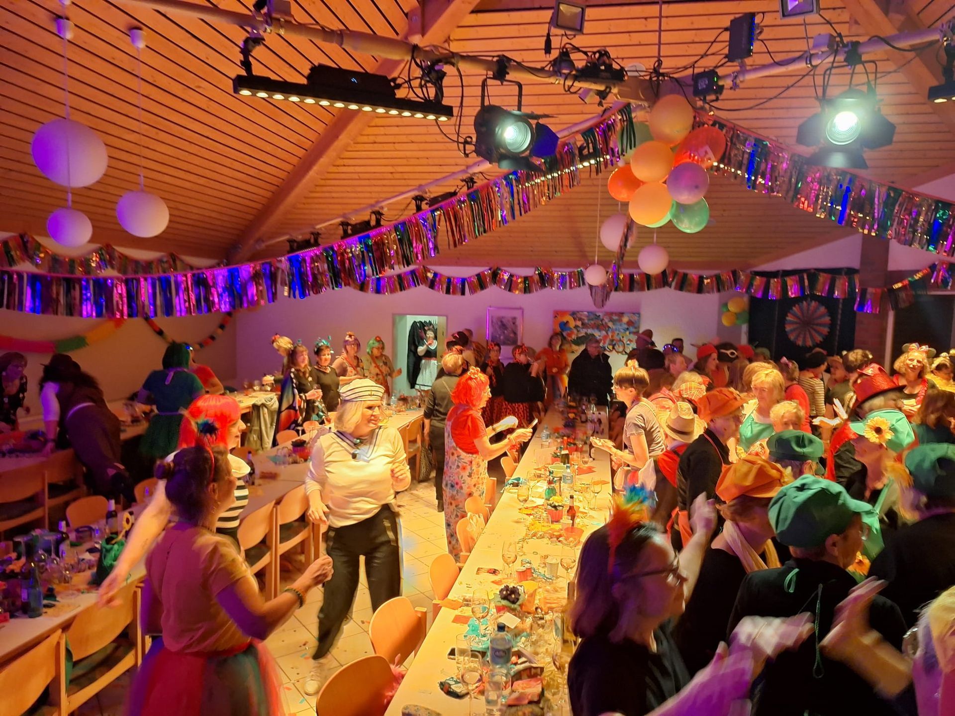 Eine große Gruppe von Menschen sitzt an langen Tischen in einem mit Luftballons dekorierten Raum.