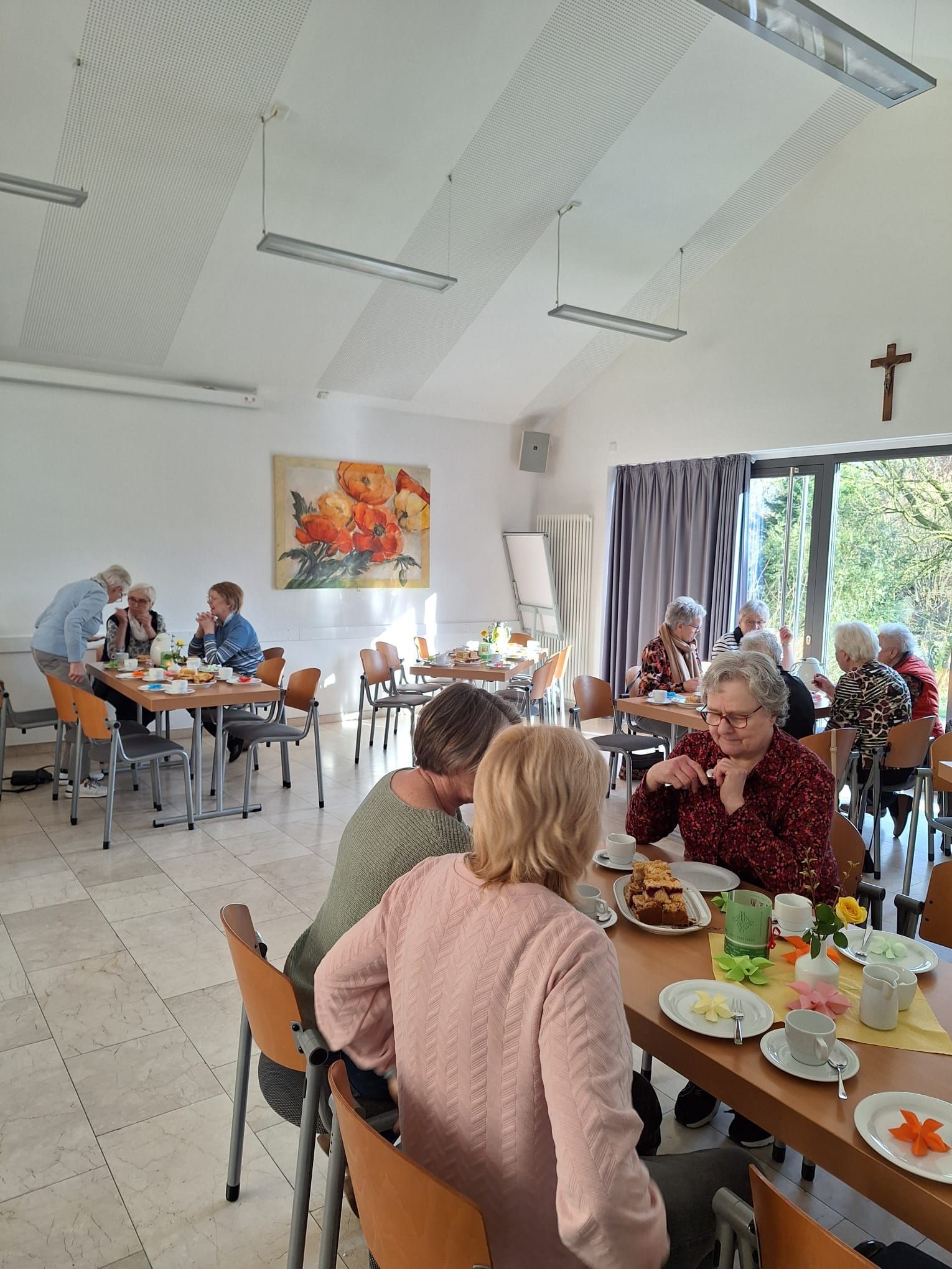 Eine Gruppe von Menschen sitzt an Tischen in einem Raum mit einem Kreuz an der Decke.