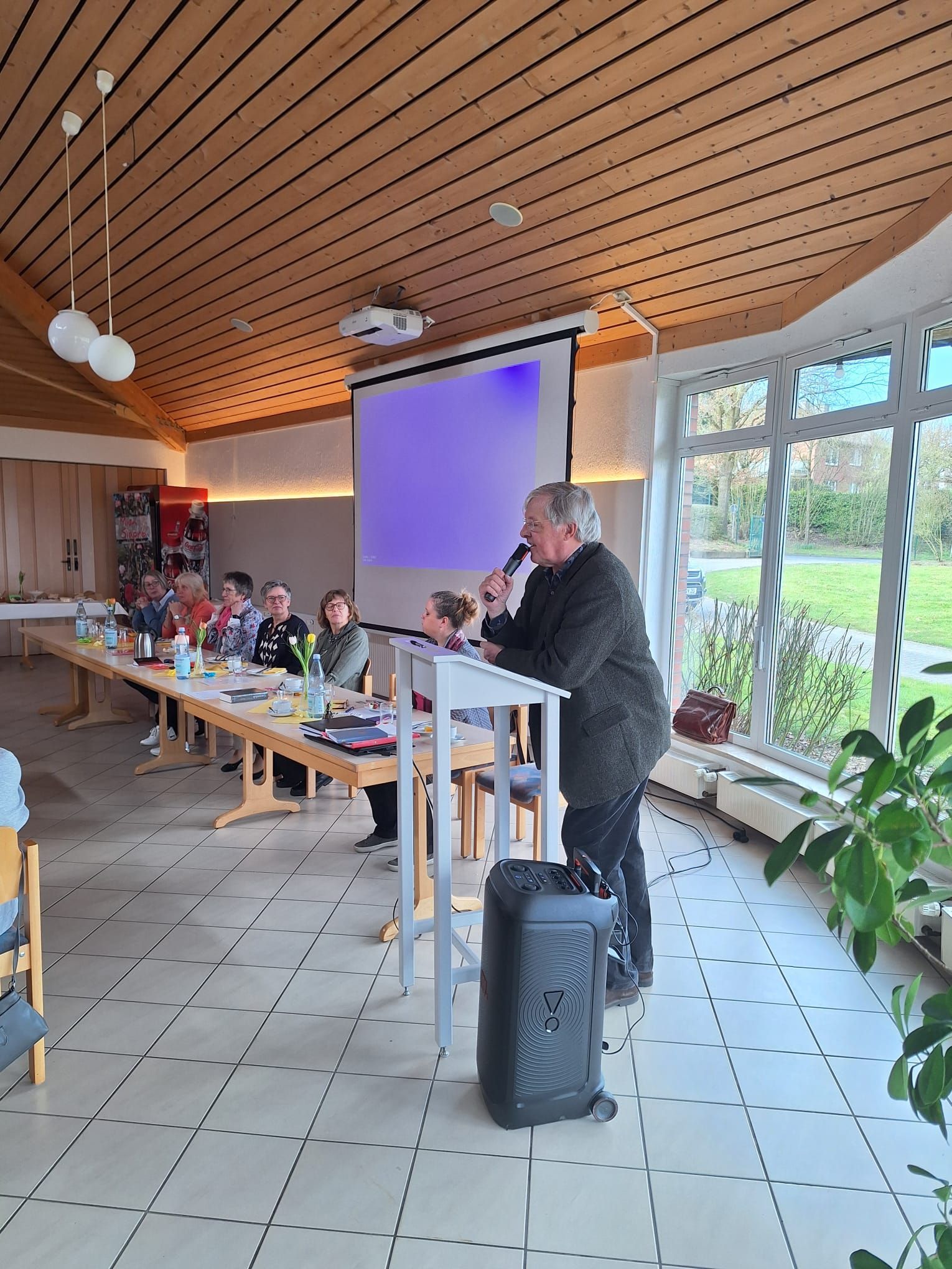 Eine Gruppe von Menschen sitzt an Tischen in einem Raum mit einer Projektionsleinwand.