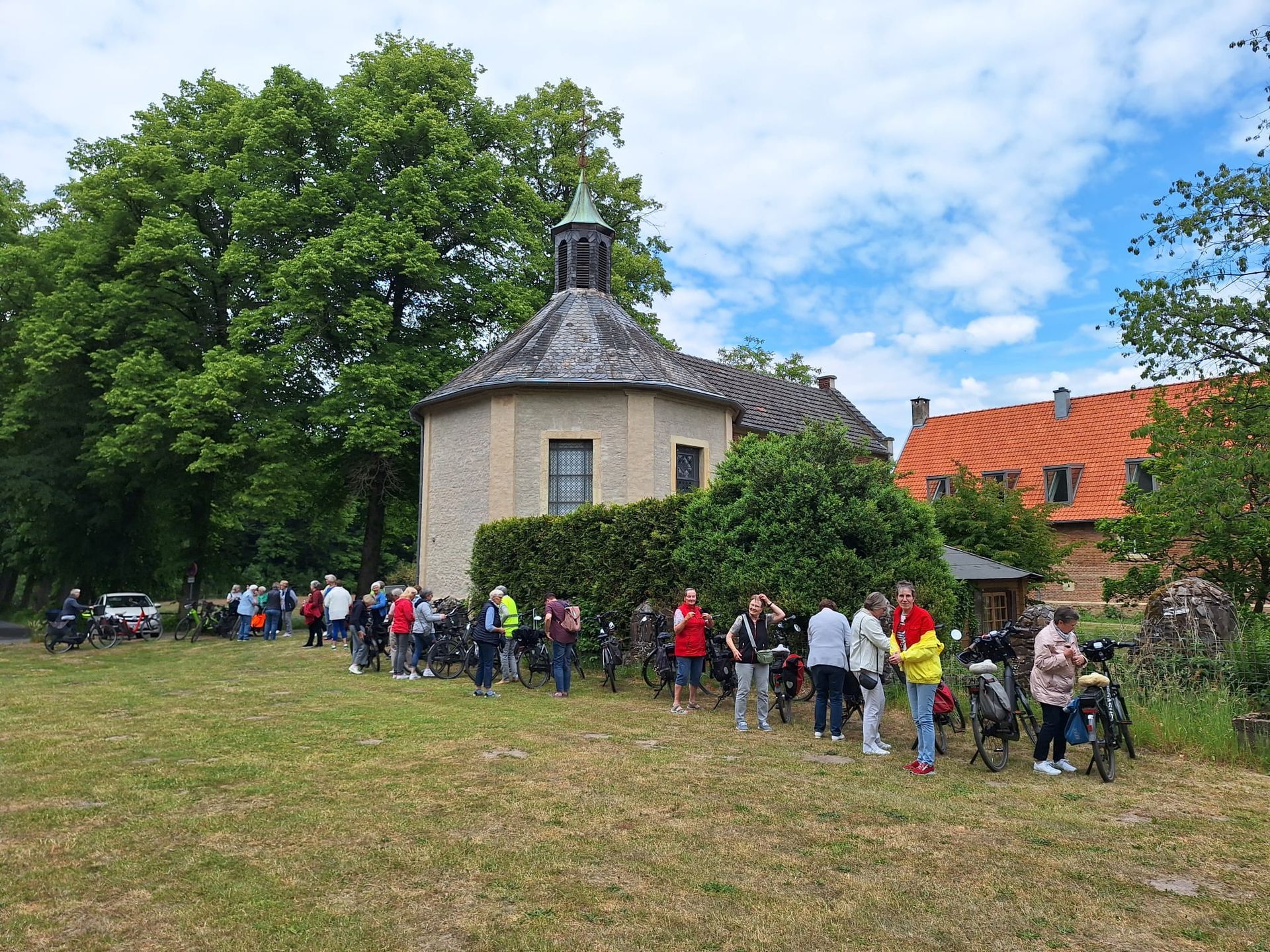 Eine Gruppe von Menschen steht auf einer Wiese vor einer kleinen Kirche.