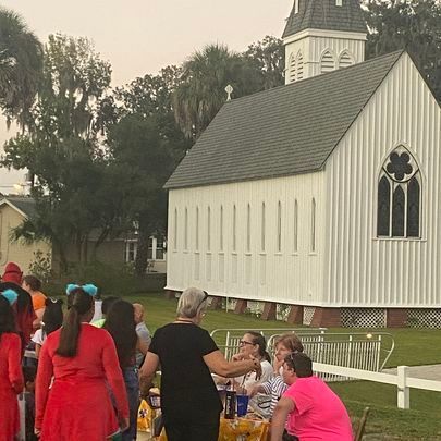 People gather outside a white church with a steeple, near a table set for a gathering.