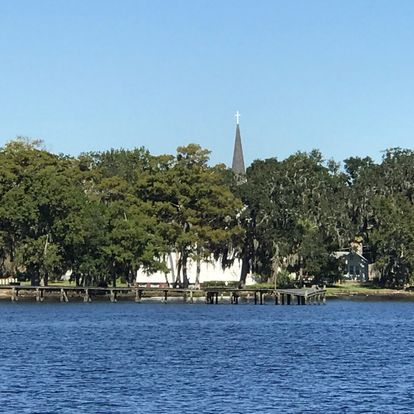 Church with steeple by water, trees, and a dock. Sunny, blue sky.