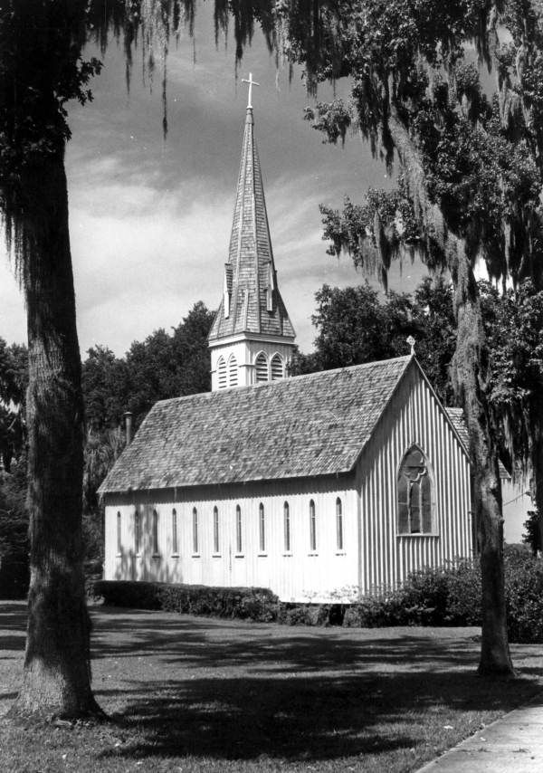 Black and white photo of a small, white church with a tall steeple, framed by trees.
