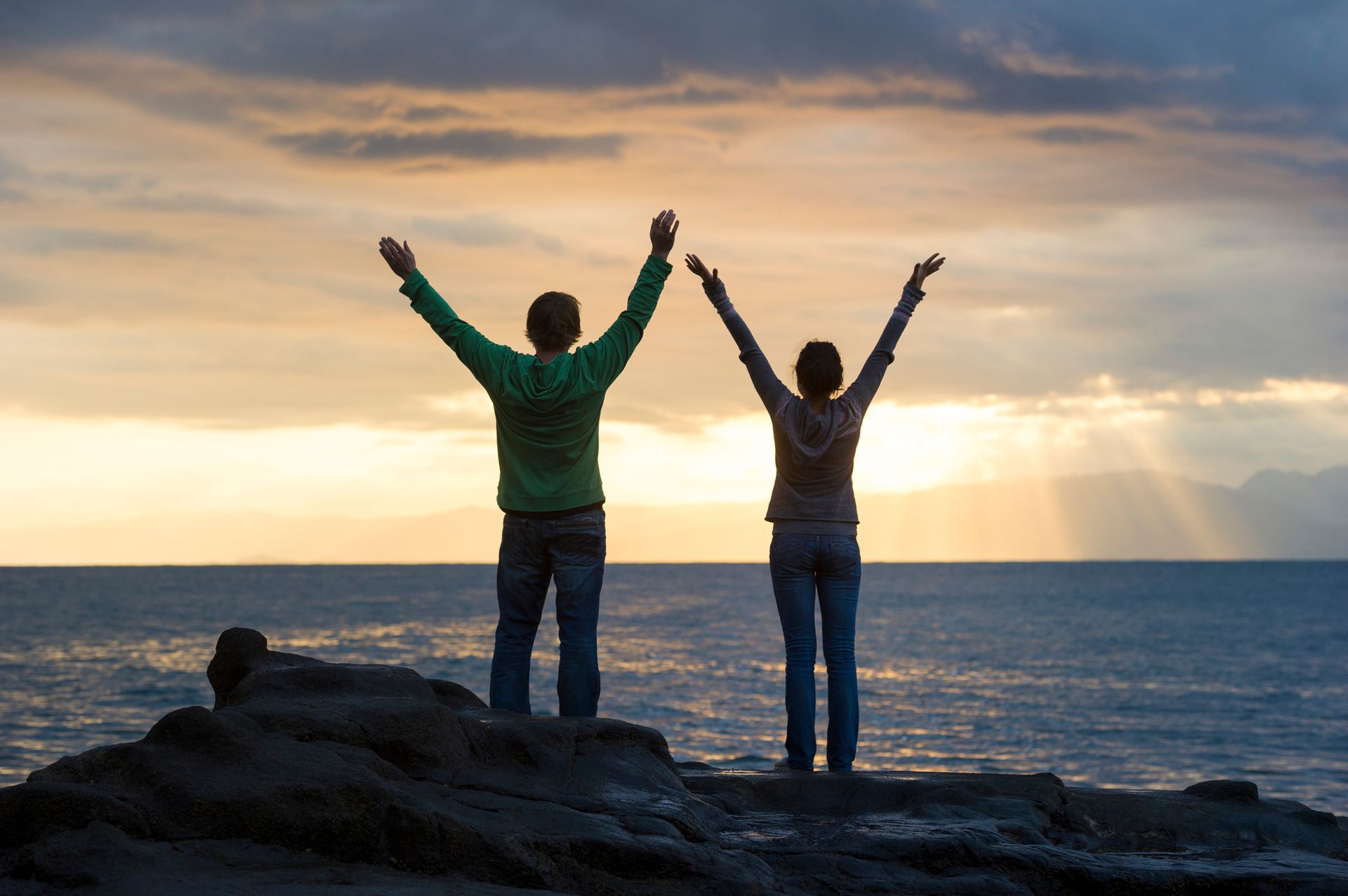 A man and a woman are standing on a rock near the ocean with their arms in the air.