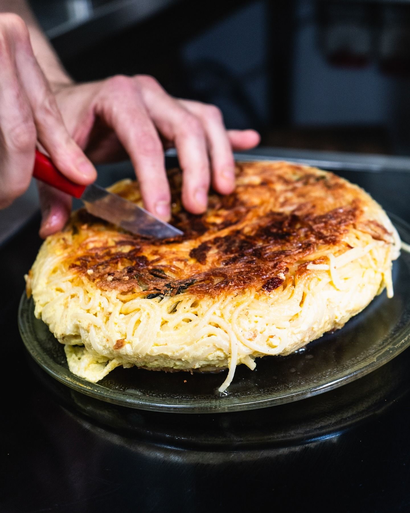 Un uomo in camice nero da chef sorride, tenendo in mano una grande palla bianca, vicino a una scatola. Bar con bottiglie.