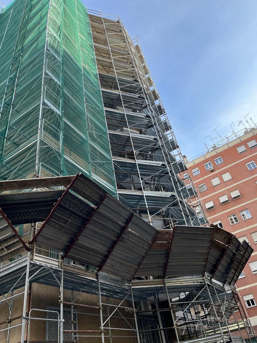 Un edificio in fase di ristrutturazione ricoperto da una rete di sicurezza verde, con un albero in primo piano e un'auto parcheggiata in strada.