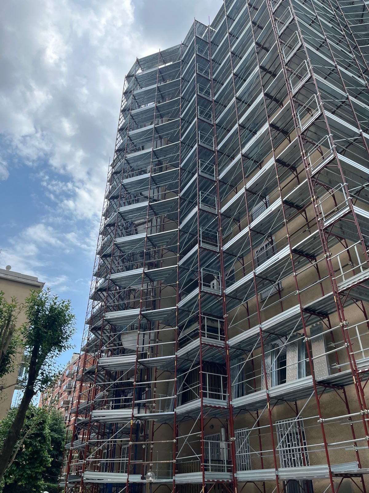 Un palazzo color beige in fase di ristrutturazione, completamente ricoperto da impalcature metalliche argentate, stagliarsi contro un cielo azzurro nuvoloso.