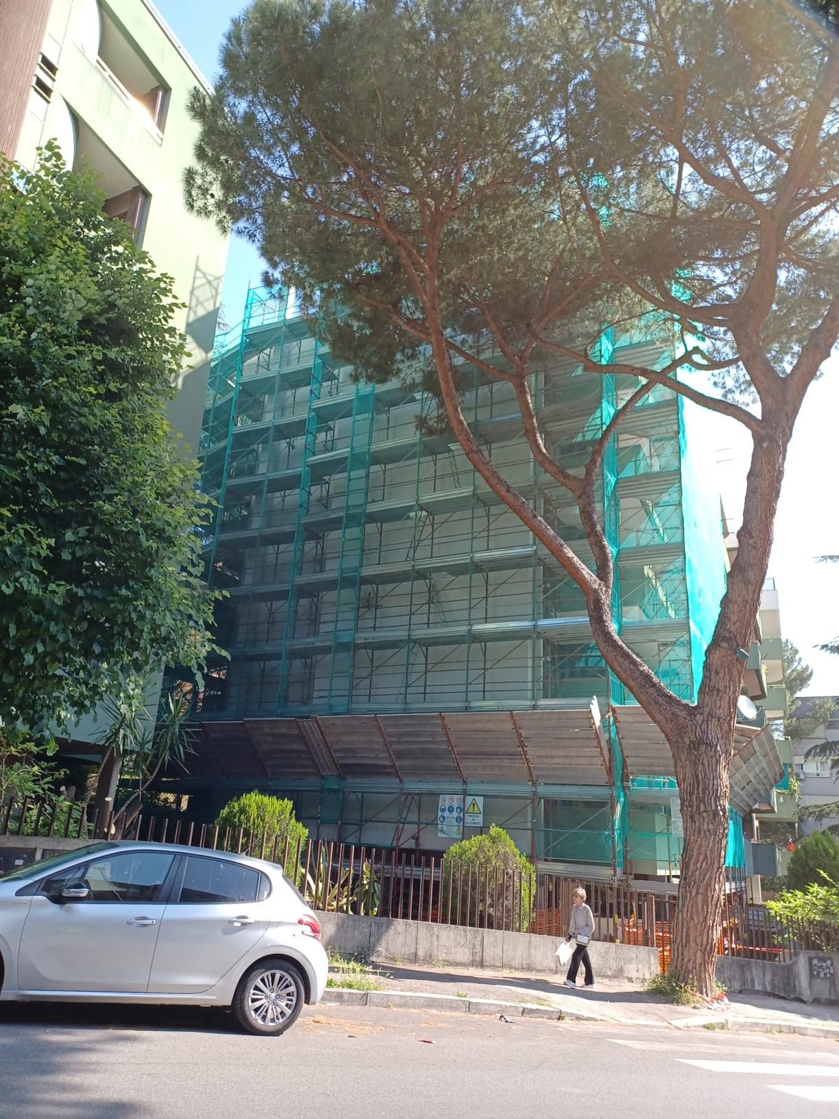 Un edificio in fase di ristrutturazione ricoperto da una rete di sicurezza verde, con un albero in primo piano e un'auto parcheggiata in strada.