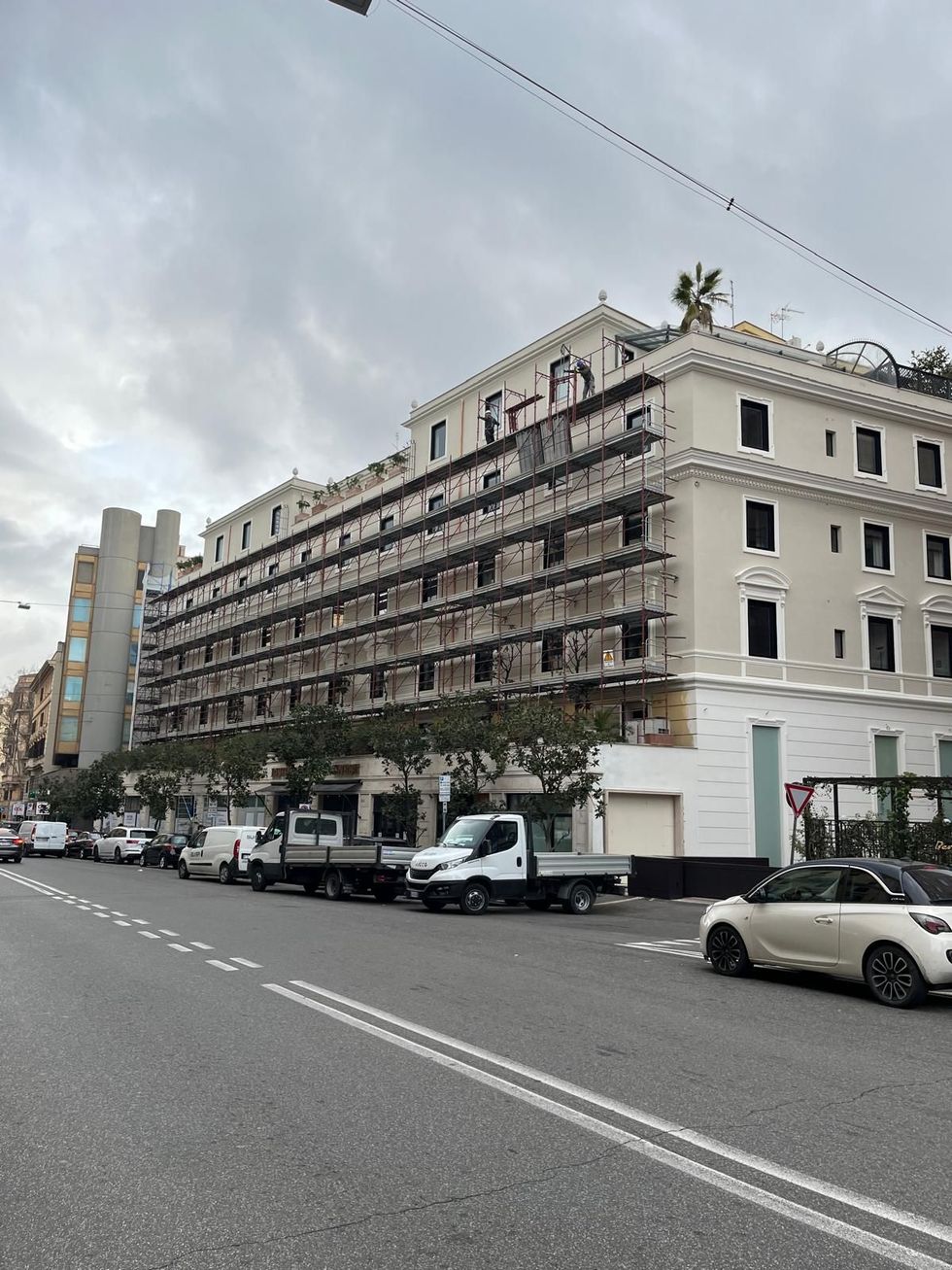 Un edificio alberghiero a più piani color crema, attualmente in fase di ristrutturazione esterna, situato su una strada con auto parcheggiate di fronte.
