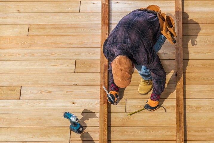 A person in work gear measuring a wooden deck board with a pencil and tape measure, with a drill resting nearby.
