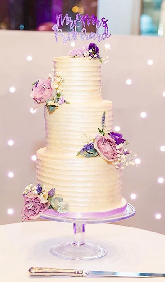 A white wedding cake with purple flowers on it is sitting on a table.