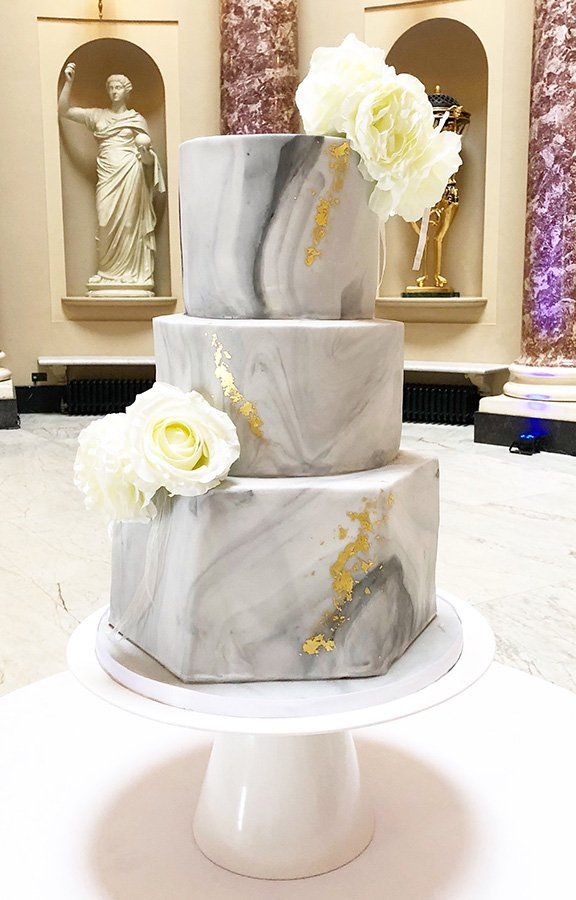 A wedding cake is sitting on top of a white cake stand in front of a statue.