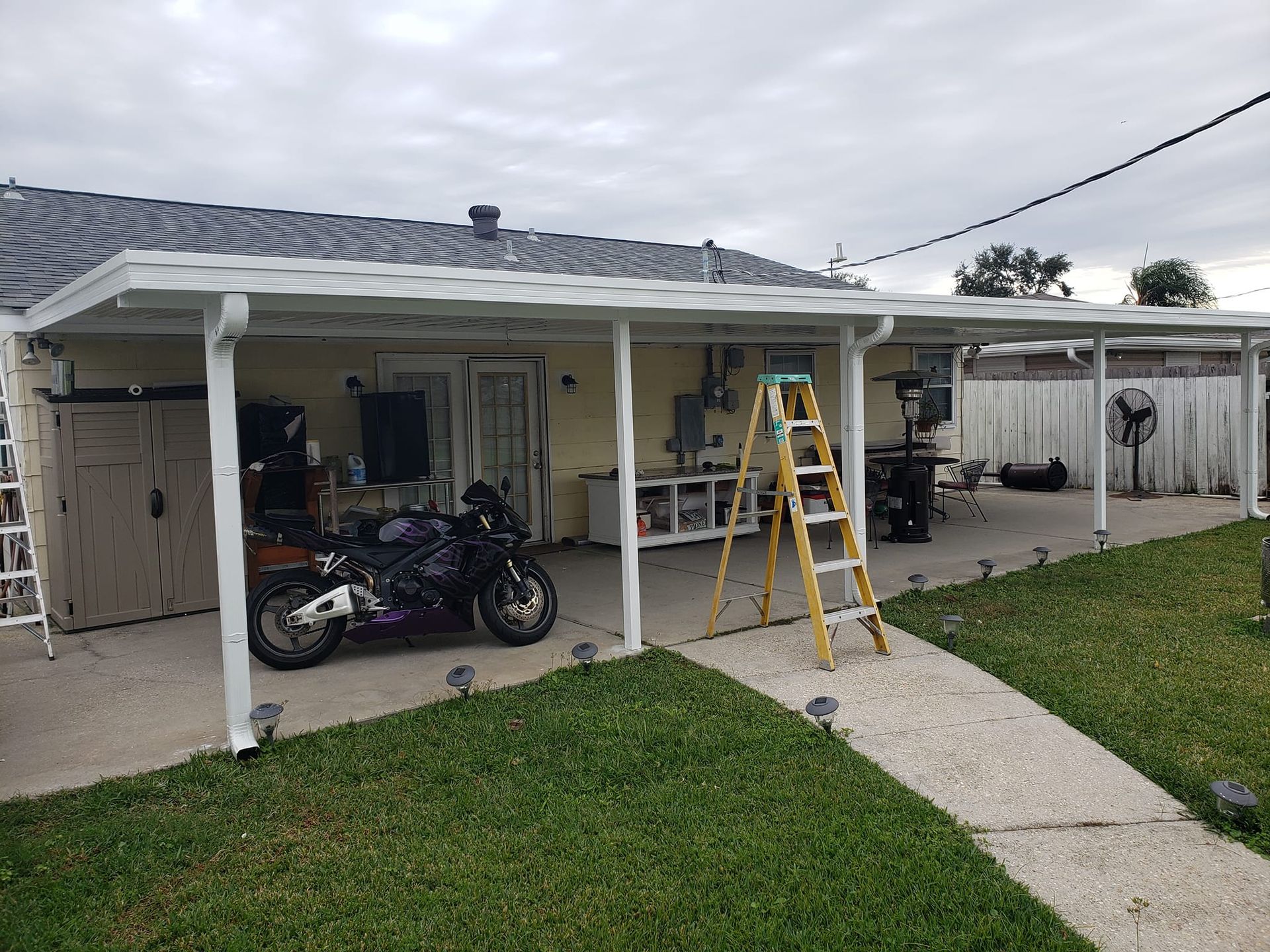 A white-roofed carport with a motorcycle, ladder, and tools, next to a yellow house on a cloudy day.