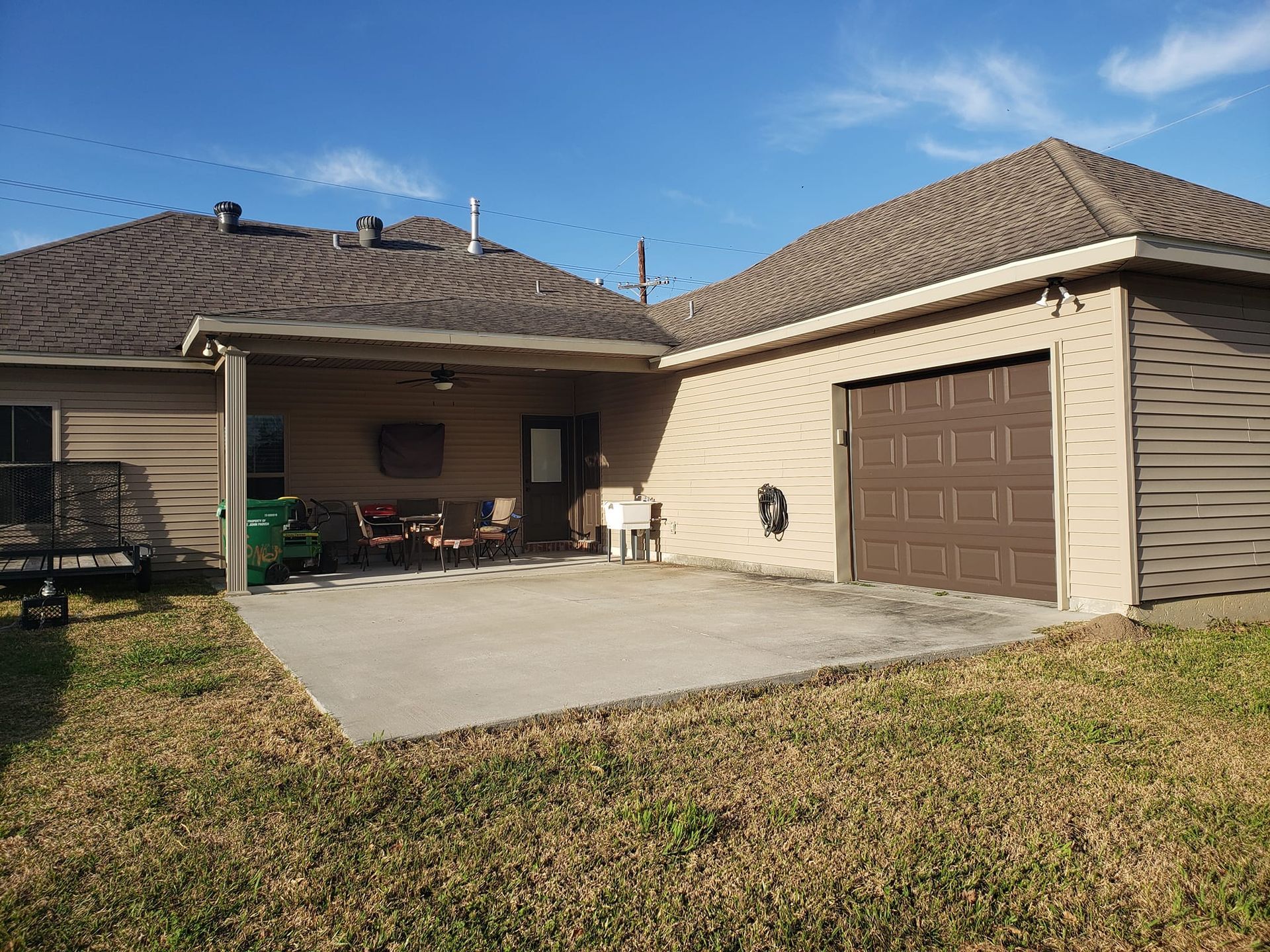 Backyard patio with brown garage and house, concrete patio, seating, and green grass. Blue sky.
