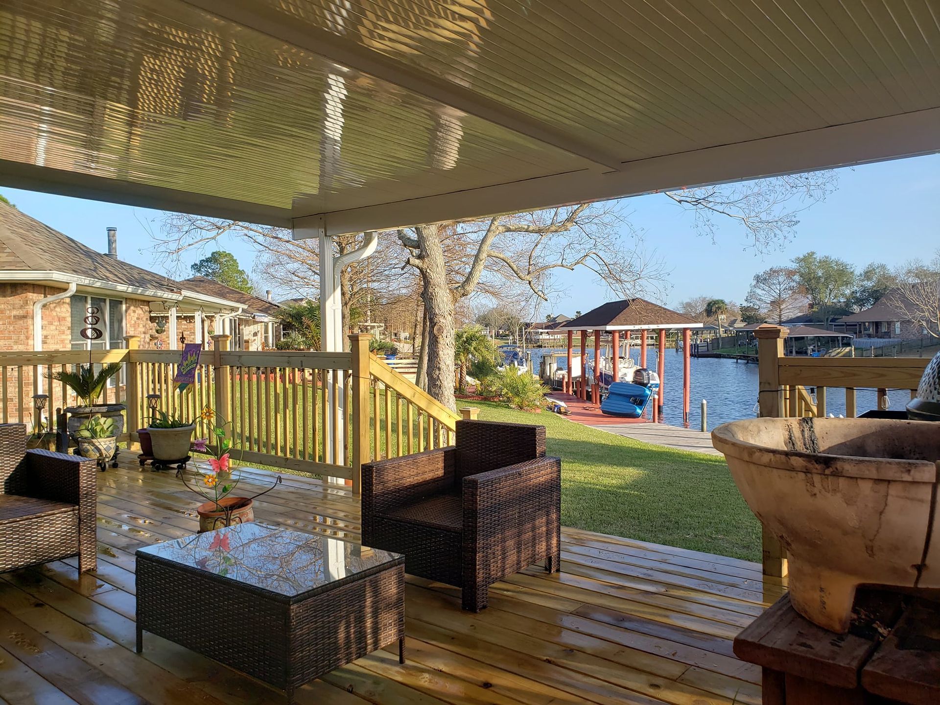 Deck with seating overlooks a lawn, gazebo, and water. Brown furniture, wooden deck, and clear sky.