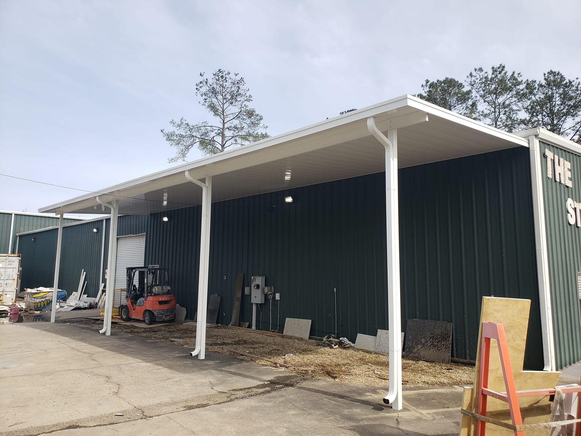 Green building with white awning, white columns, and a forklift on a concrete surface.