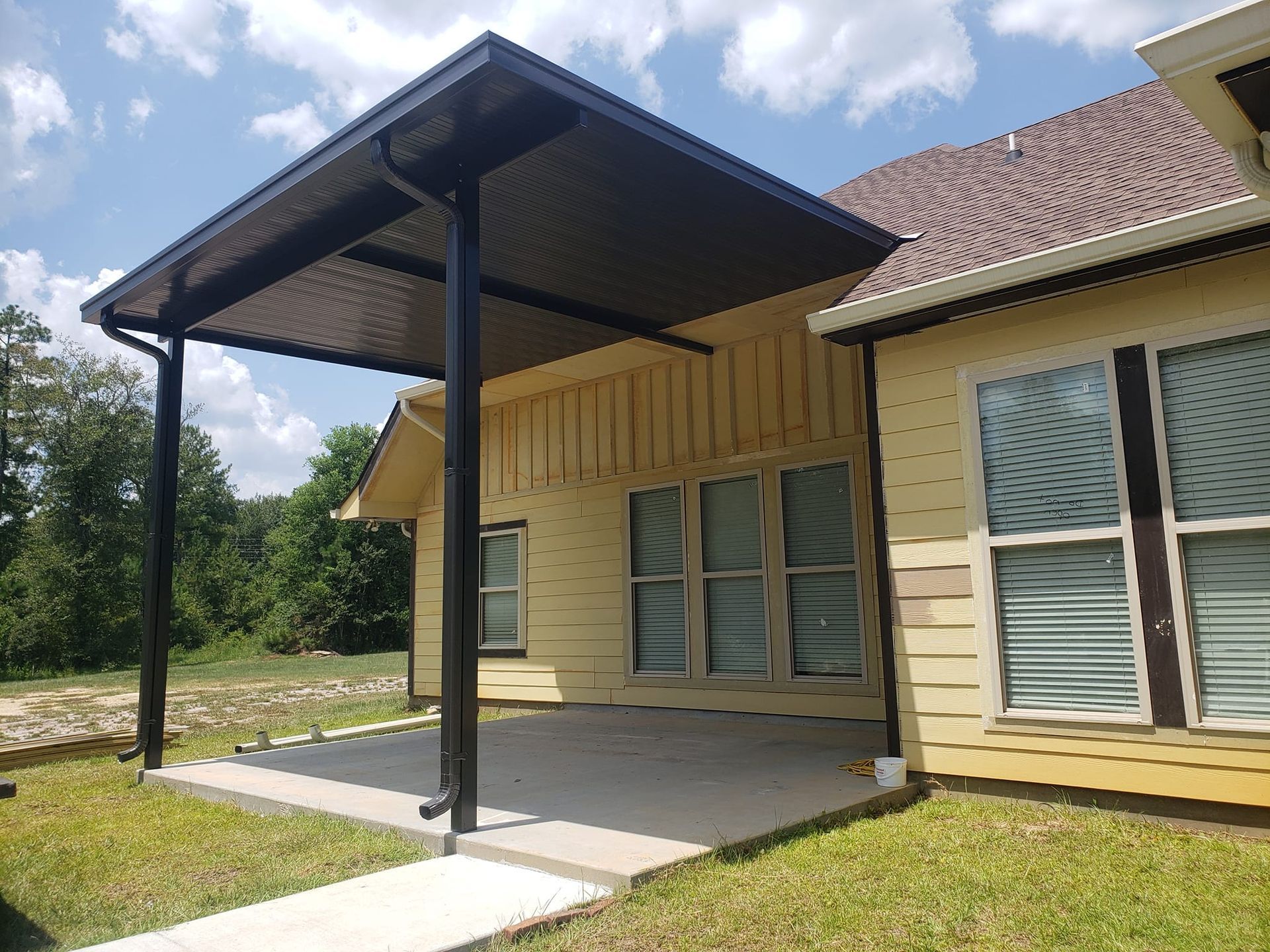 Black pergola attached to a yellow house, over a concrete patio. Green grass surrounds.