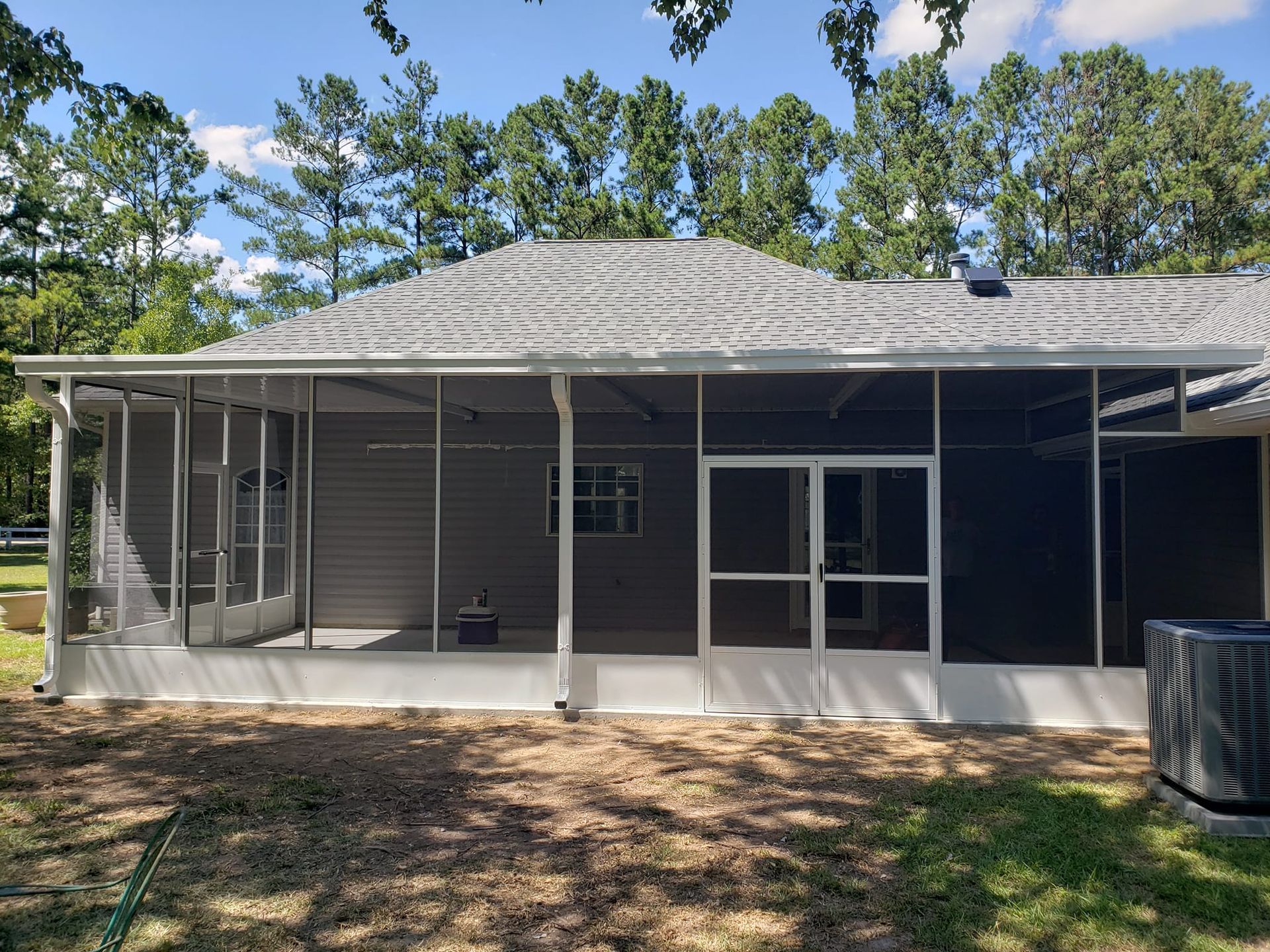 Screened-in porch attached to a gray house, with white trim, and surrounded by grass and trees.