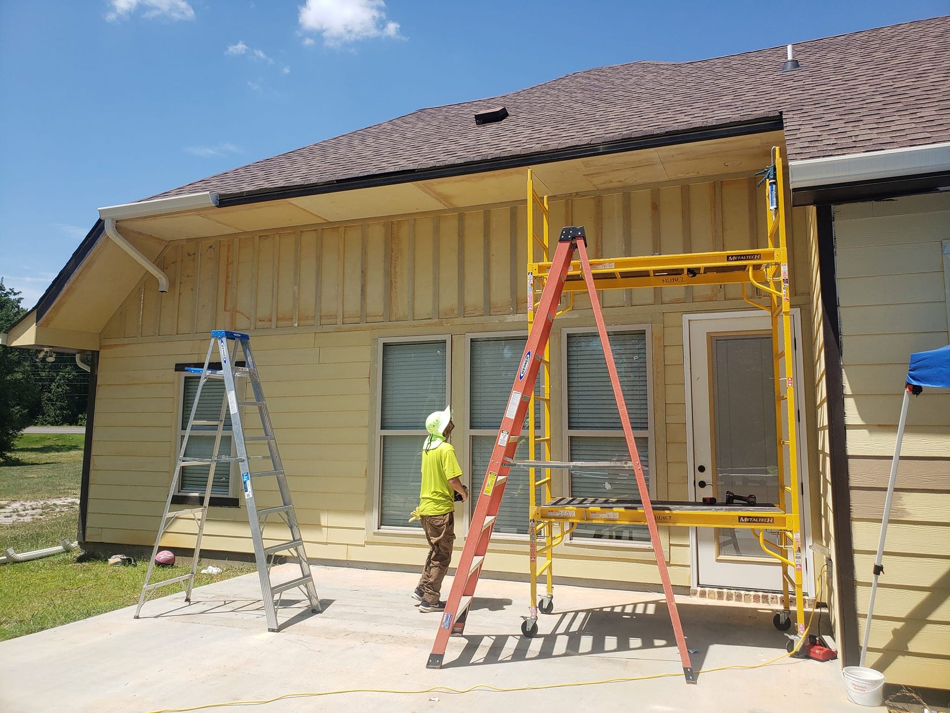 Person in yellow shirt on a scaffold painting a yellow house, with a ladder to the left, on a sunny day.