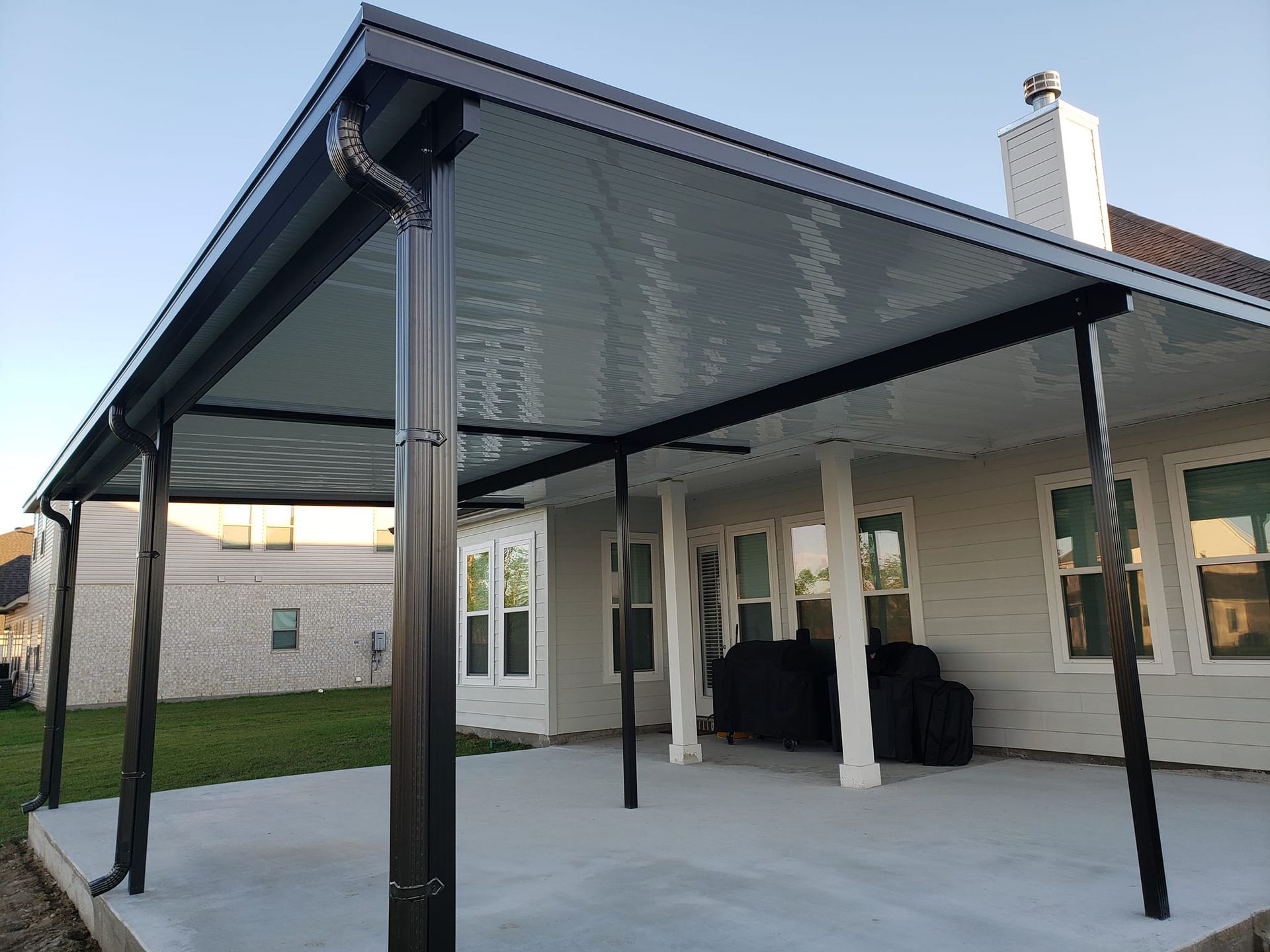 Black patio cover attached to a house with a concrete patio, green lawn, and blue sky.