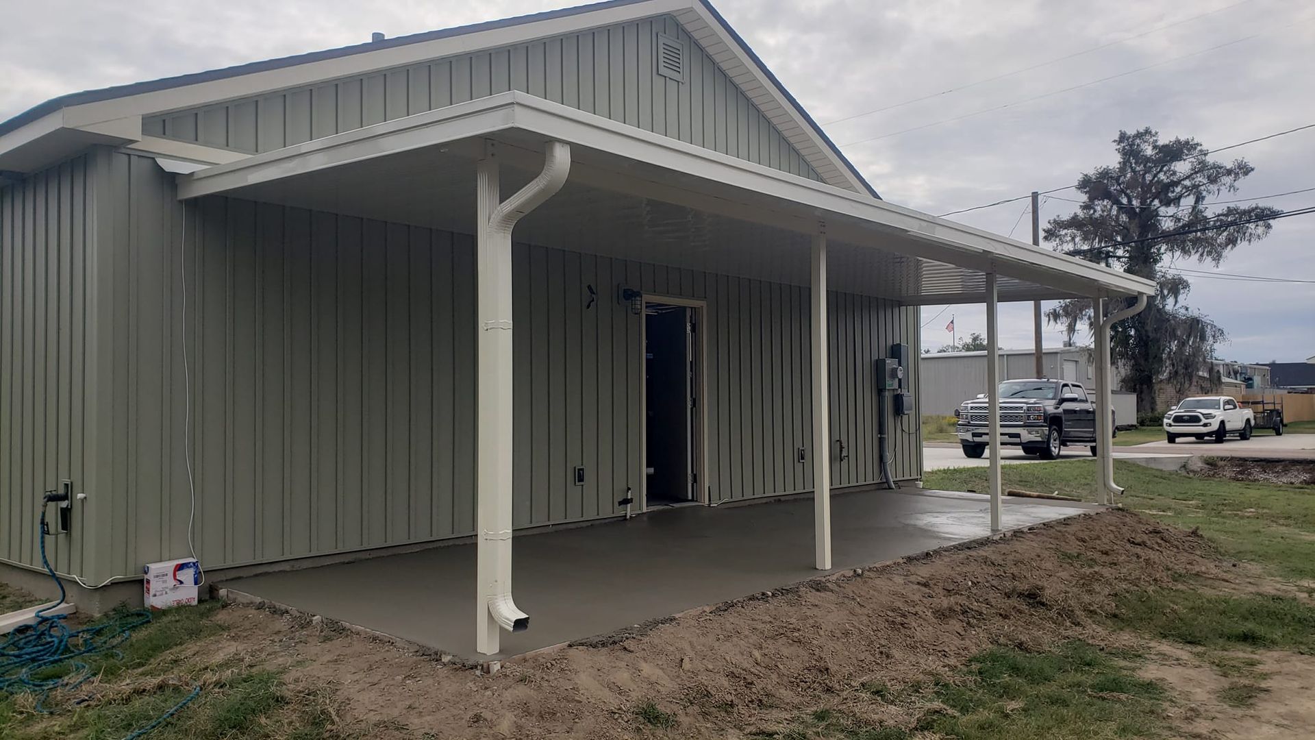 Green building with a concrete patio and covered entryway, with a few vehicles visible in the background.