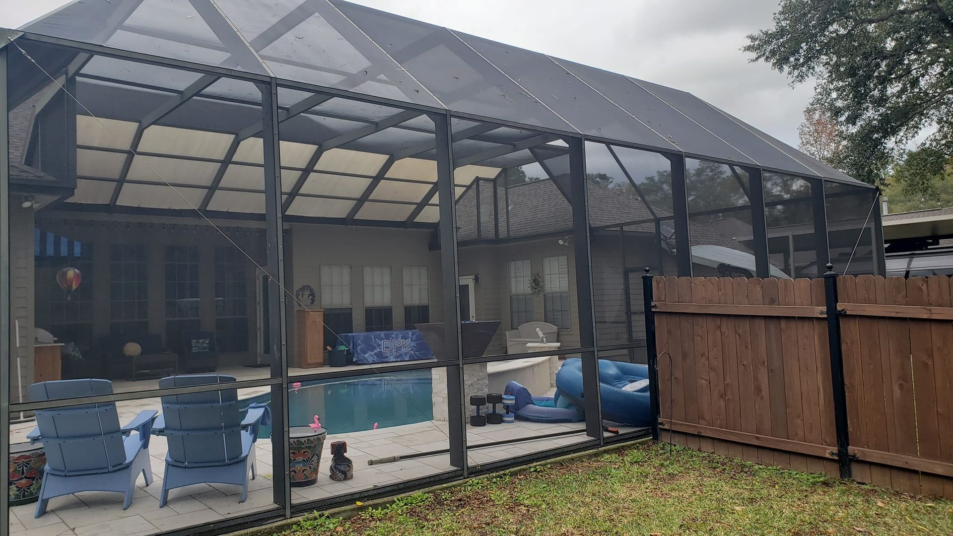 A screened-in pool enclosure with blue chairs, pool, and patio area next to a wooden fence.
