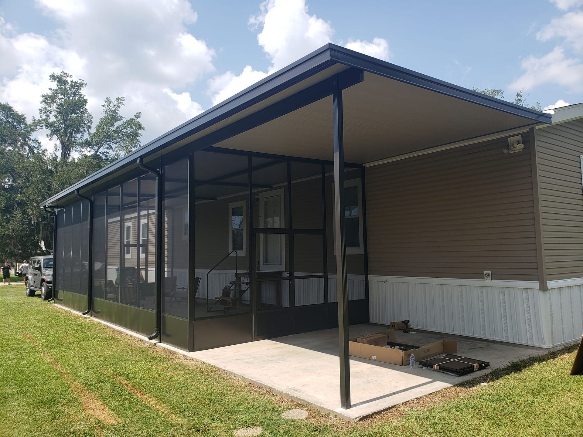 Screened-in porch attached to a mobile home with a black frame and tan roof; green yard and blue sky.