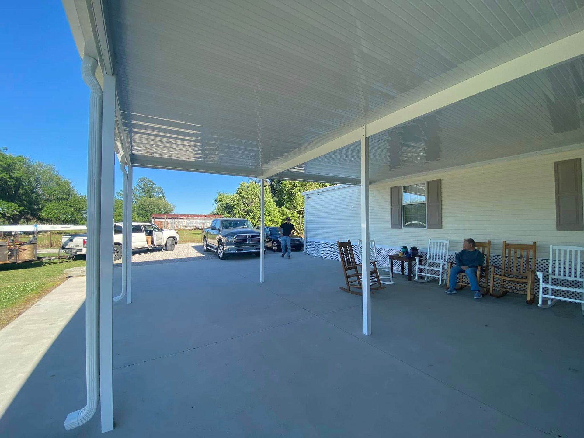 Covered patio with people, vehicles, and rocking chairs. Blue sky, white columns, gray floor.