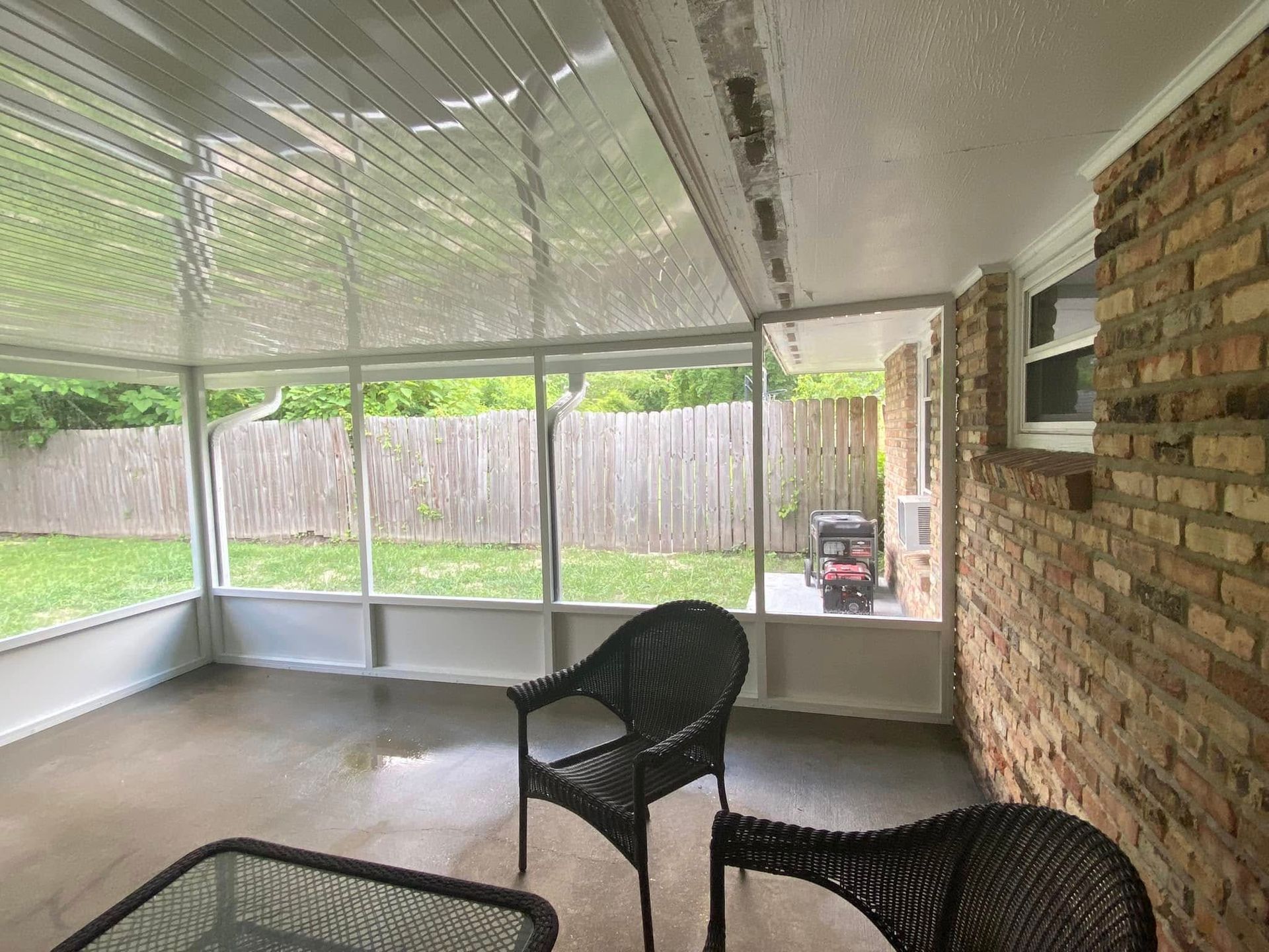 Screened-in patio with two wicker chairs, a table, and a view of a yard. Brick wall to the right.