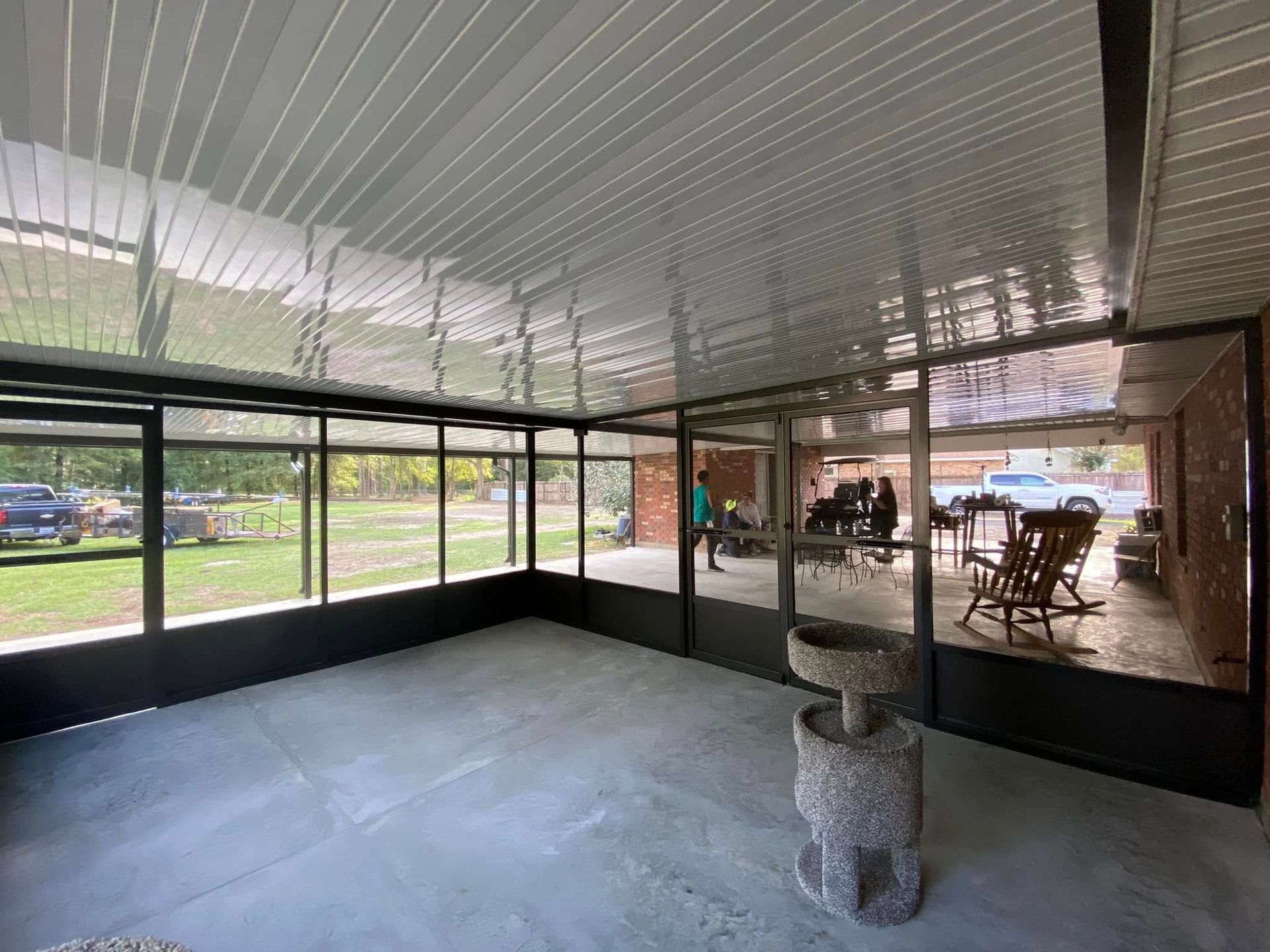 Sunroom with concrete floor, enclosed with black frames and screens, attached to a house. People are visible in the background.