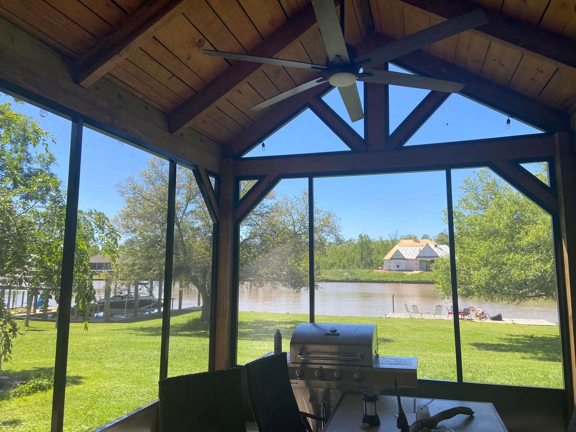 Screened-in porch with view of a lake, trees, and a house. Grill and table are in the foreground.