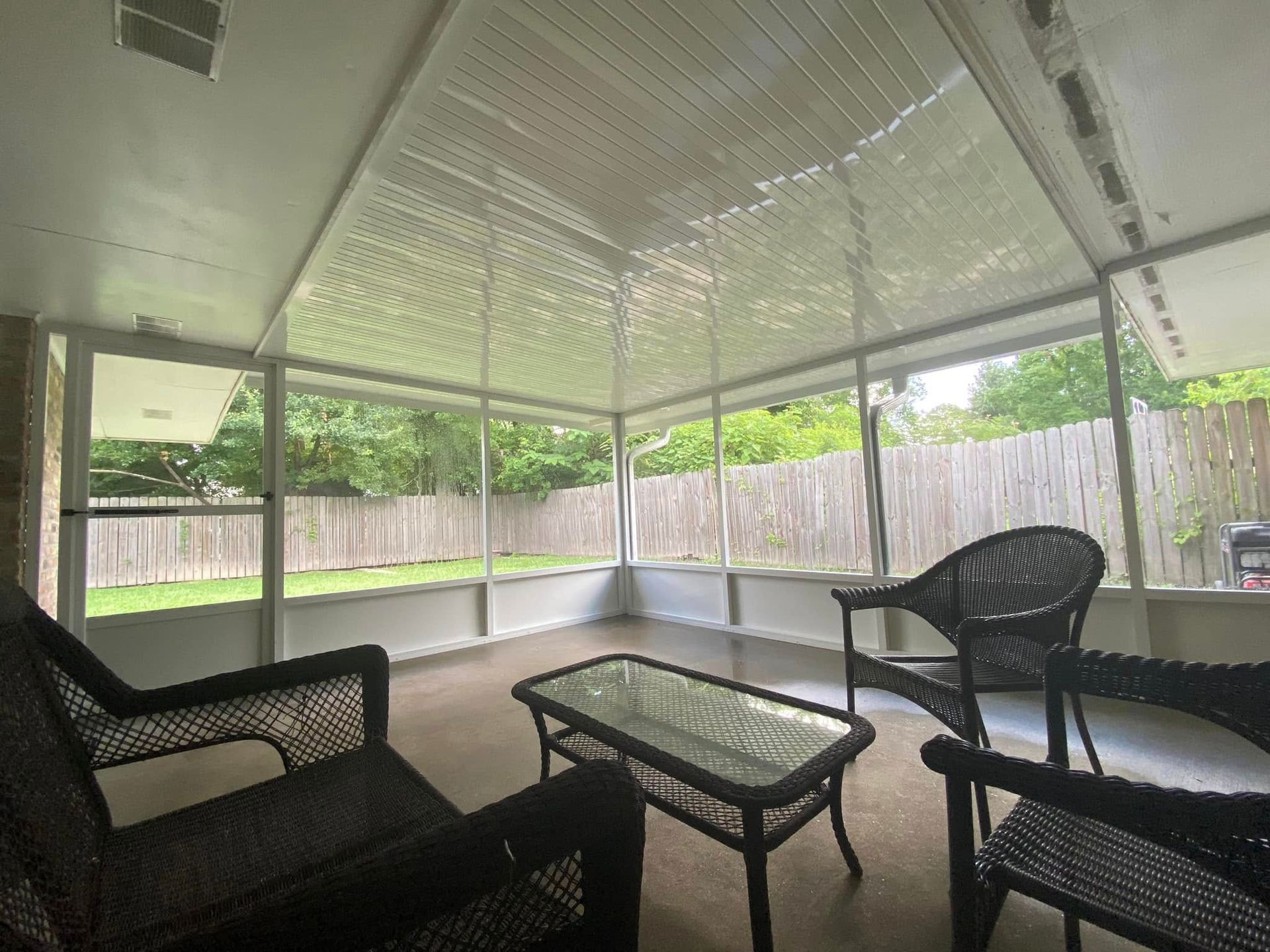 Sunroom with wicker furniture, glass table, and screened windows overlooking a wooded backyard.