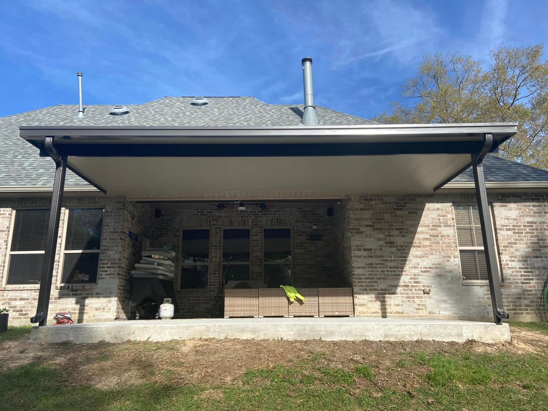 A covered patio with black supports and a white awning, attached to a brick house under a blue sky.