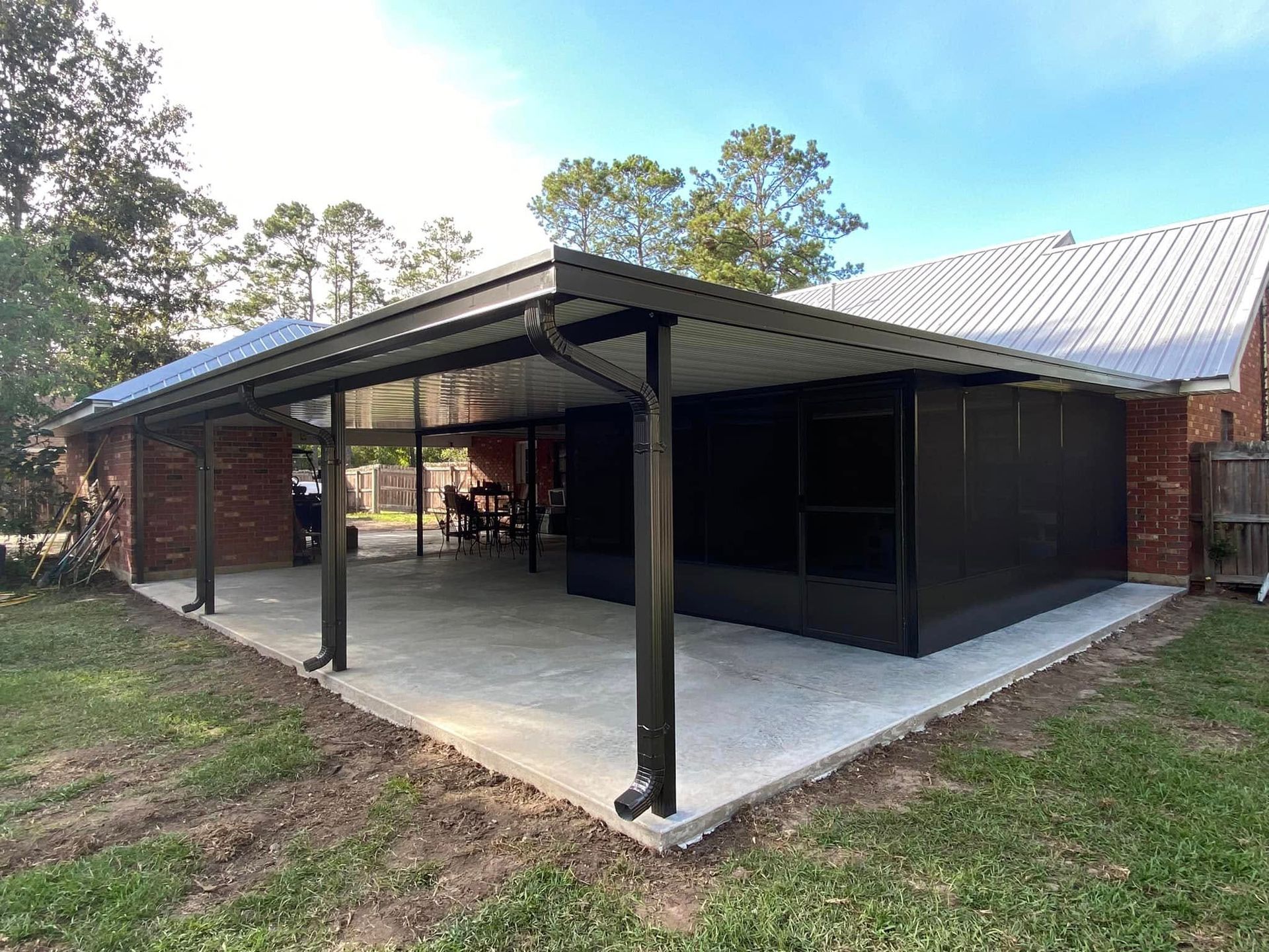 A patio with a black roof and support beams, attached to a brick house, on a concrete slab.